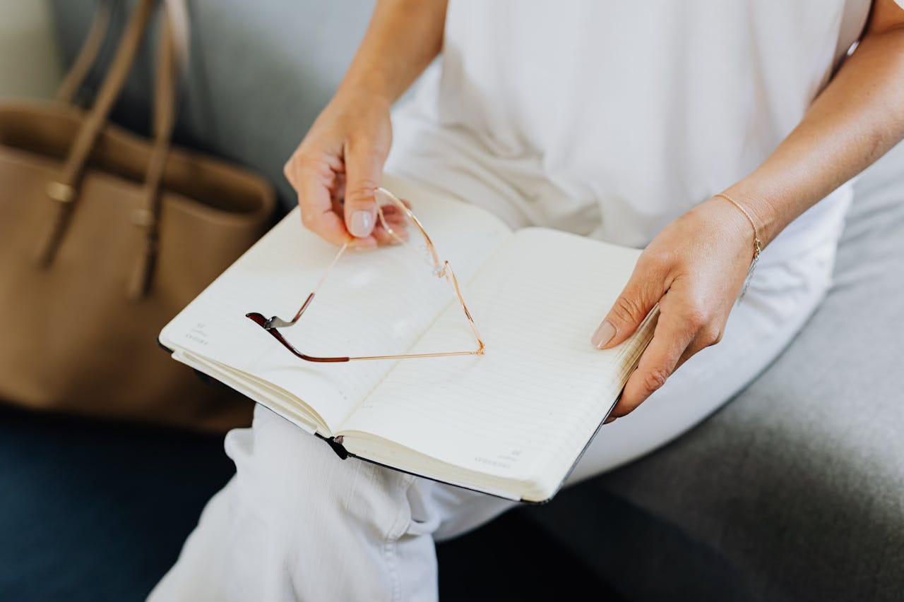 Woman in all white sits on a stool holding her glasses in one hand and a journal sits on her knee.