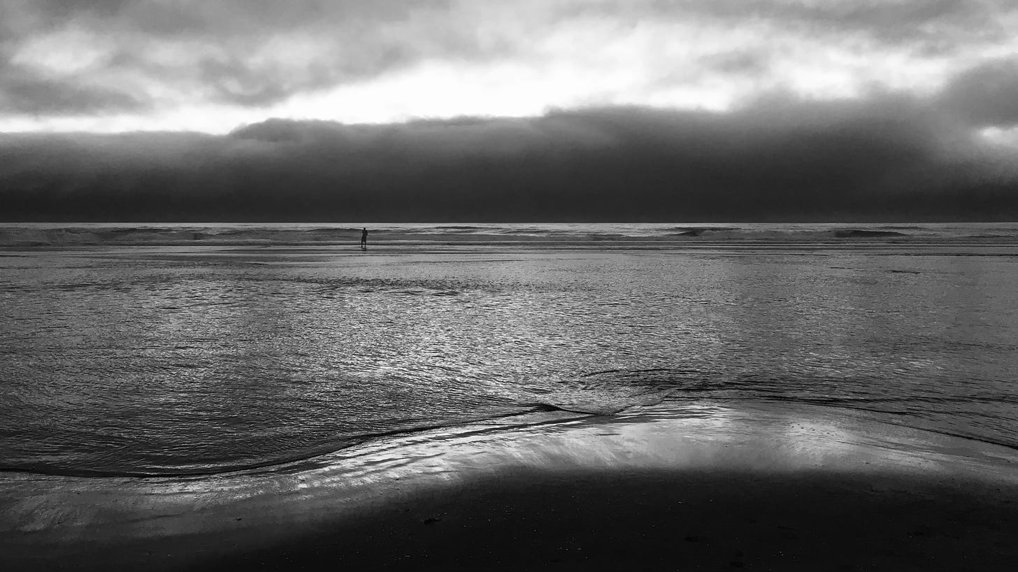 Black-and-white photo of an ocean beach shortly after sunset, with fog on the horizon and a man standing in very shallow water in the distance.