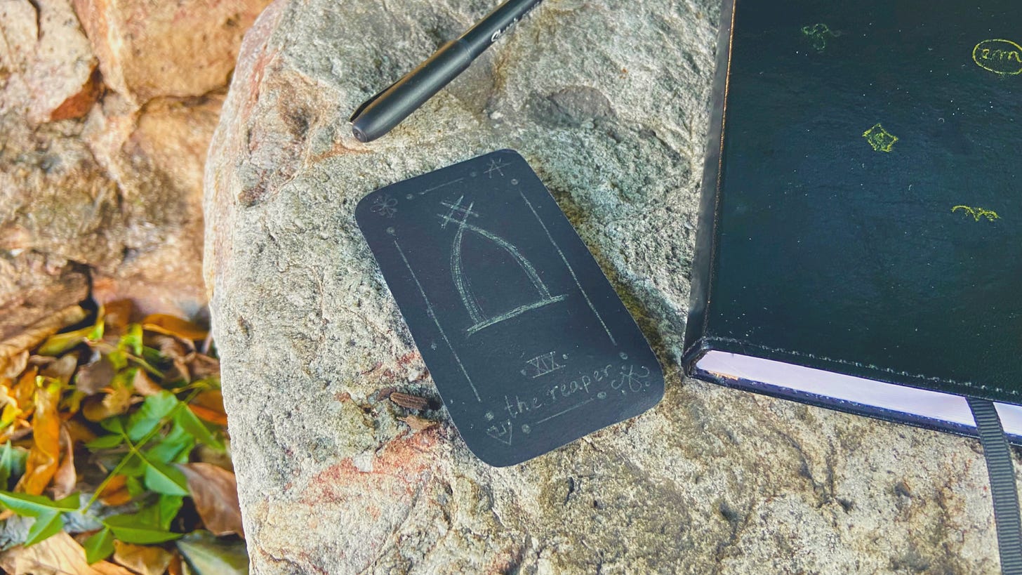 A black oracle card with silver writing that depicts an abstract headstone and the words "the reaper". The card sits on a rock next to a black journal with a pen beside it. There are beginnings of autumn leaves on the ground beneath the rock.