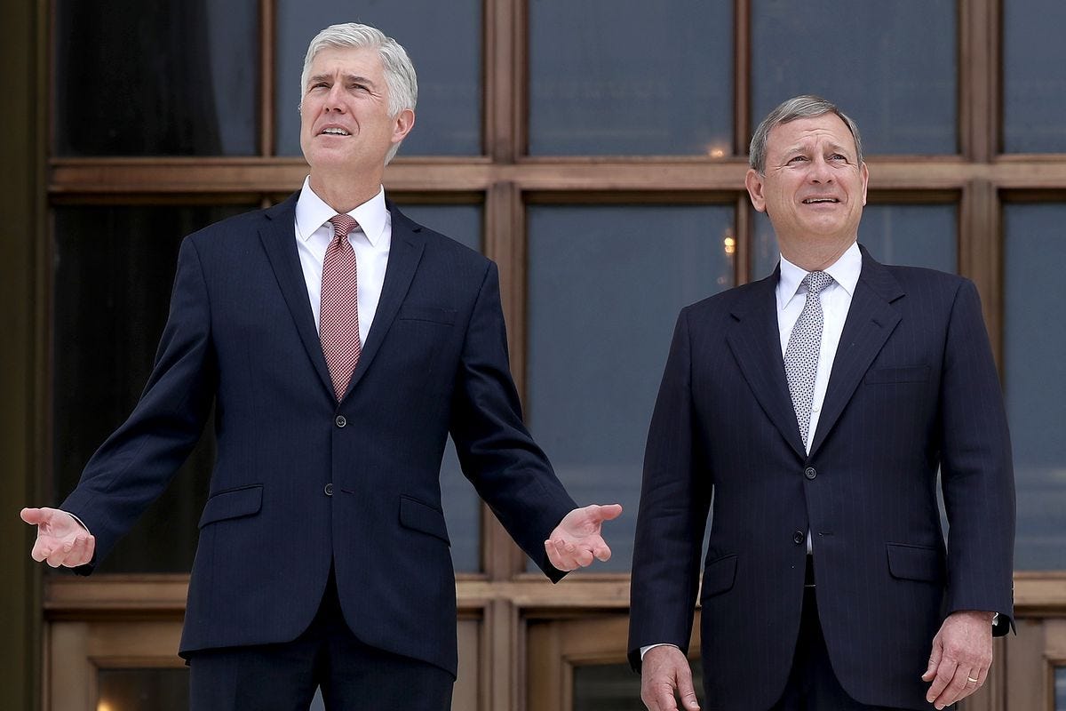 Justice Neil Gorsuch, left, in a navy suit and red tie, and Chief Justice John Roberts, right, in a black suit and gray tie, stand in front of the Supreme Court building.