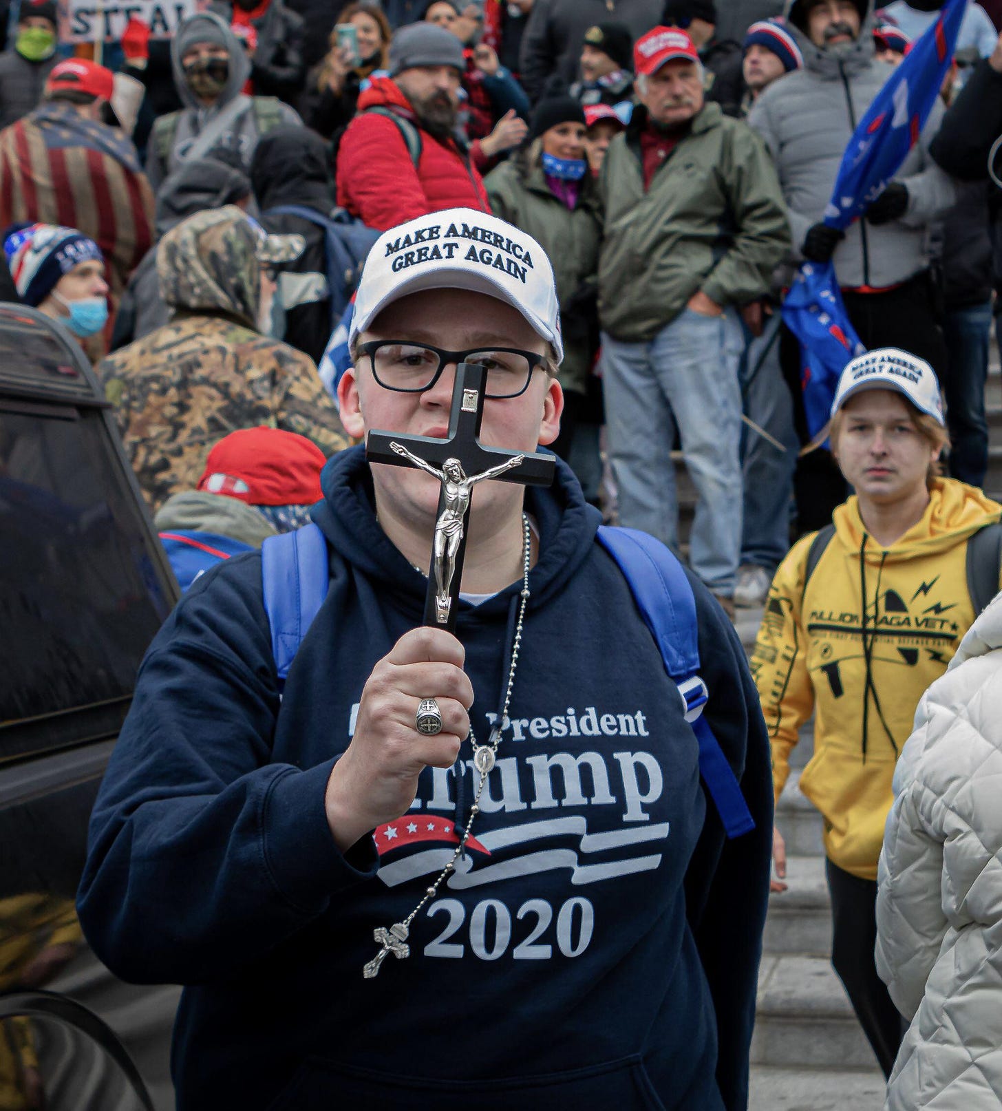 A man wearing a MAGA hat and Trump 2020 sweatshirt is holding up a crucifix