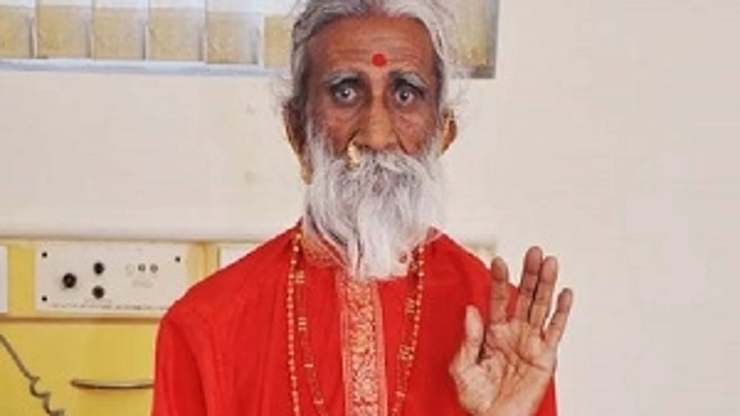 El asceta Prahlad Jani mira de frente la cámara y levanta su  mano izquierda a modo de saludo.