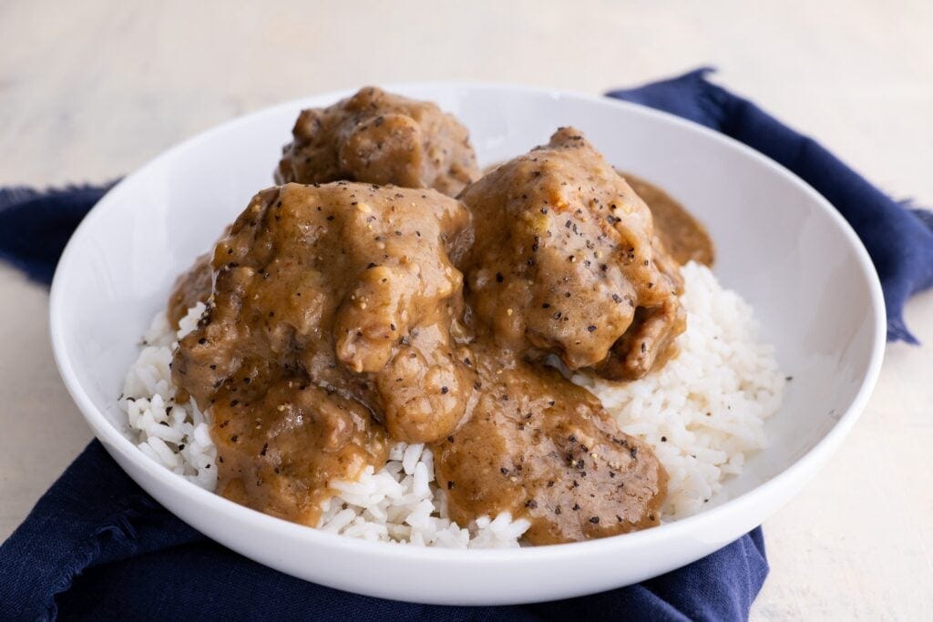 a white bowl of oxtails covered in brown gravy over a bed of rice and a blue napkin in the background