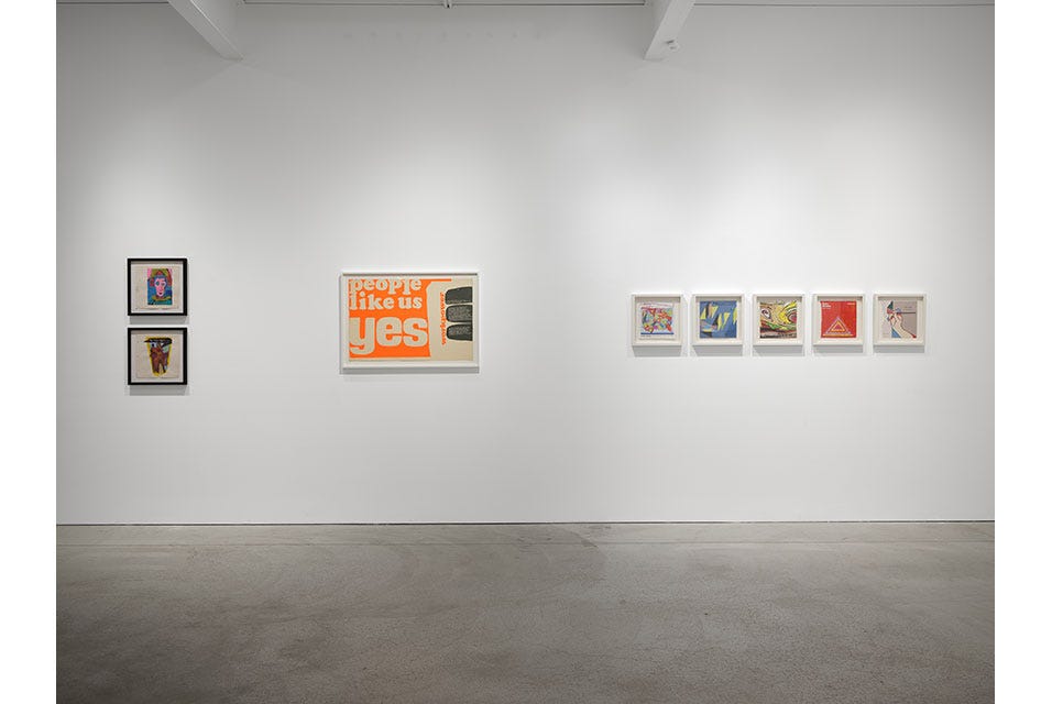 A photo of a gallery with a grey floor and brightly lit white walls. Several frames artworks hang on the wall. One of them has a bright orange background with text that says, people like us, yes.