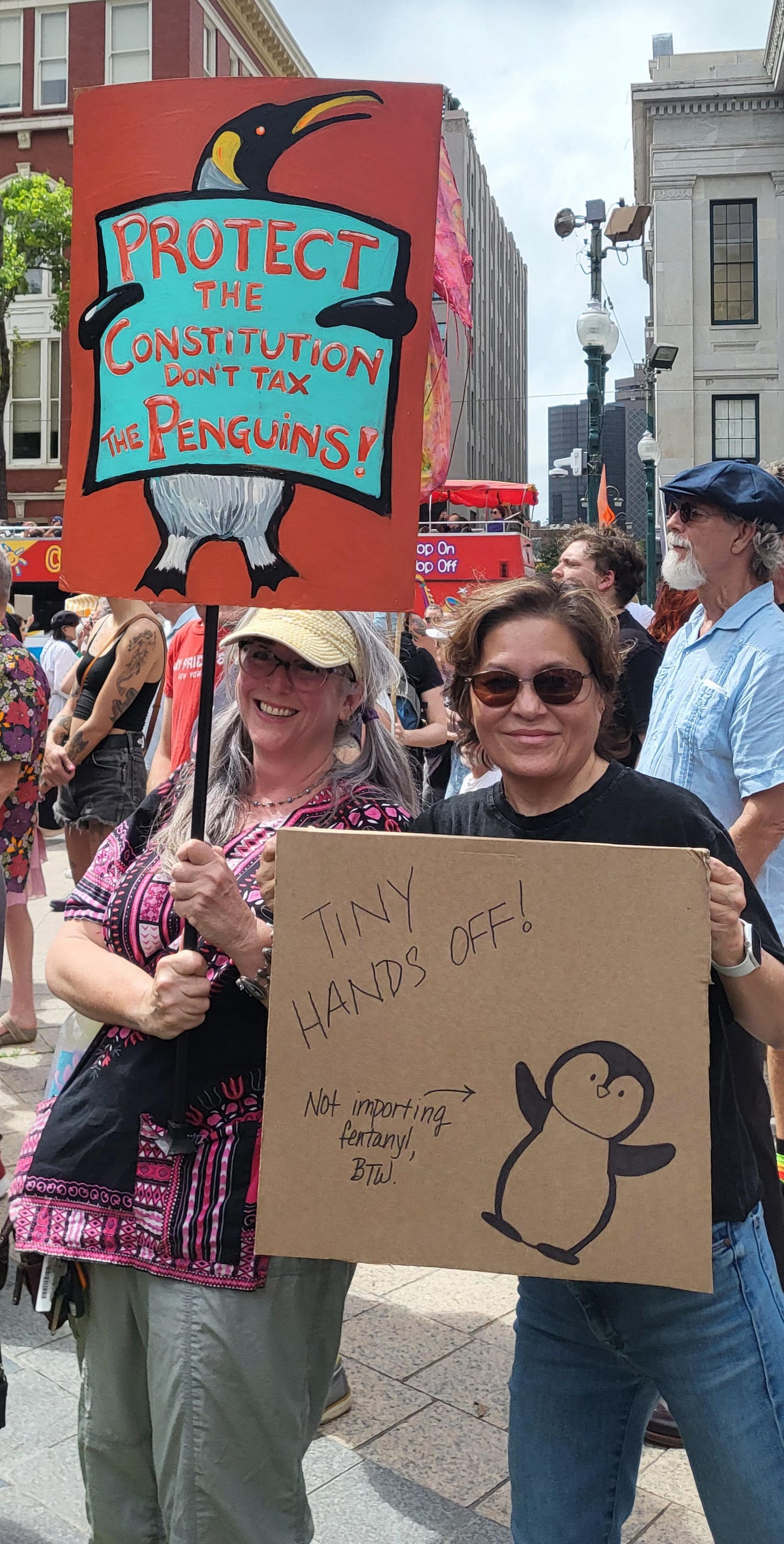 A group of protesters holding signs on a New Orleans sidewalk. One sign is red, with a painting of a penguin holding a sign that reads "Protect the Constitution, Don't Tax the Penguins." The other is a cardboard sign with a drawing of a penguin labeled "not importing fentanyl" and "Tiny Hands Off" A group of protesters holding signs on a New Orleans sidewalk. One sign is red, with a painting of a penguin holding a sign that reads "Protect the Constitution, Don't Tax the Penguins." The other is a cardboard sign with a drawing of a penguin labeled "not importing fentanyl" and "Tiny Hands Off"