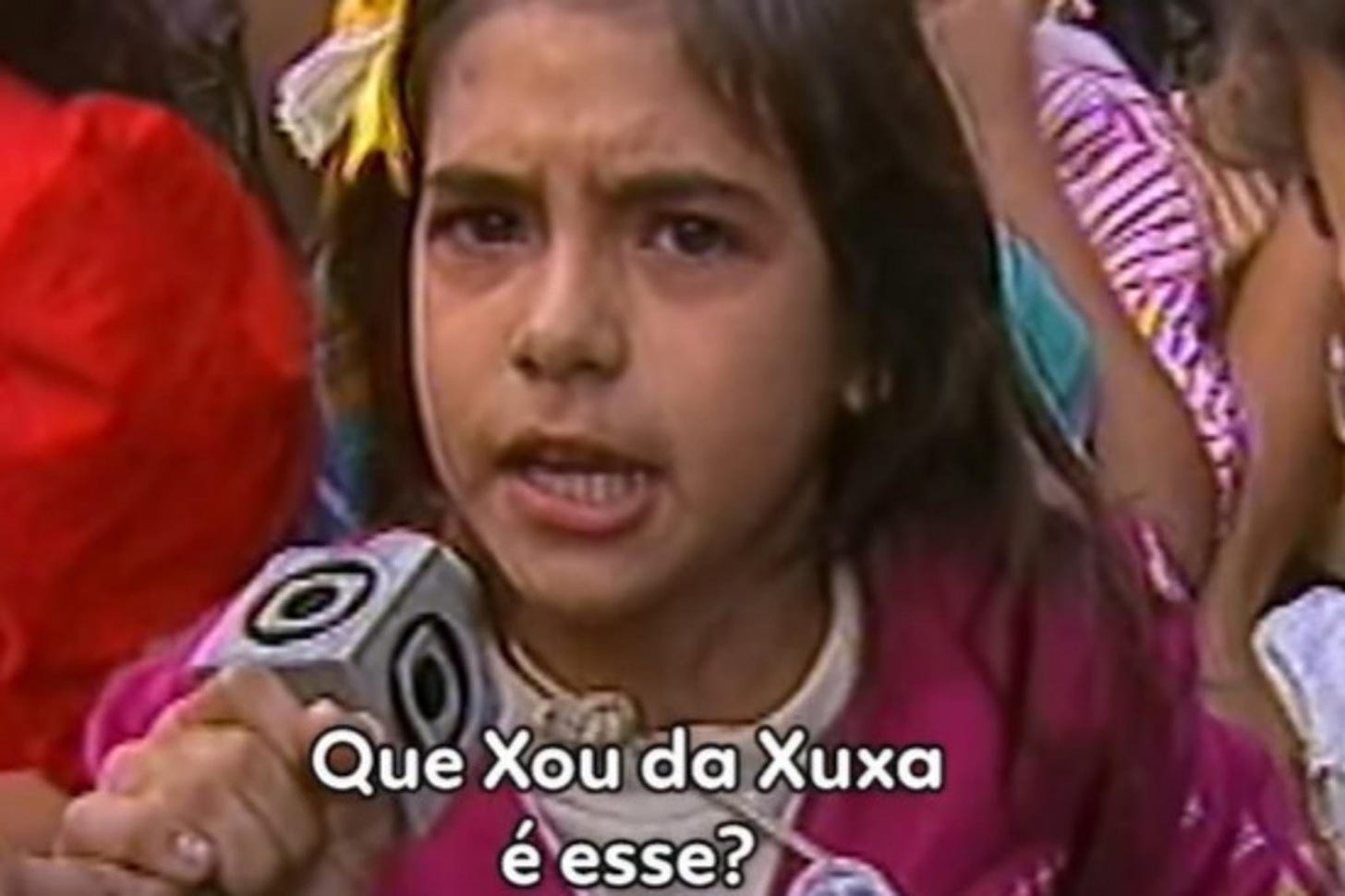 Uma menina de uns 8, 9 anos, com laço da cabeça e blusa rosa, segurando o microfone do repórter da globo enquanto pergunta "que show da Xuxa é esse?"