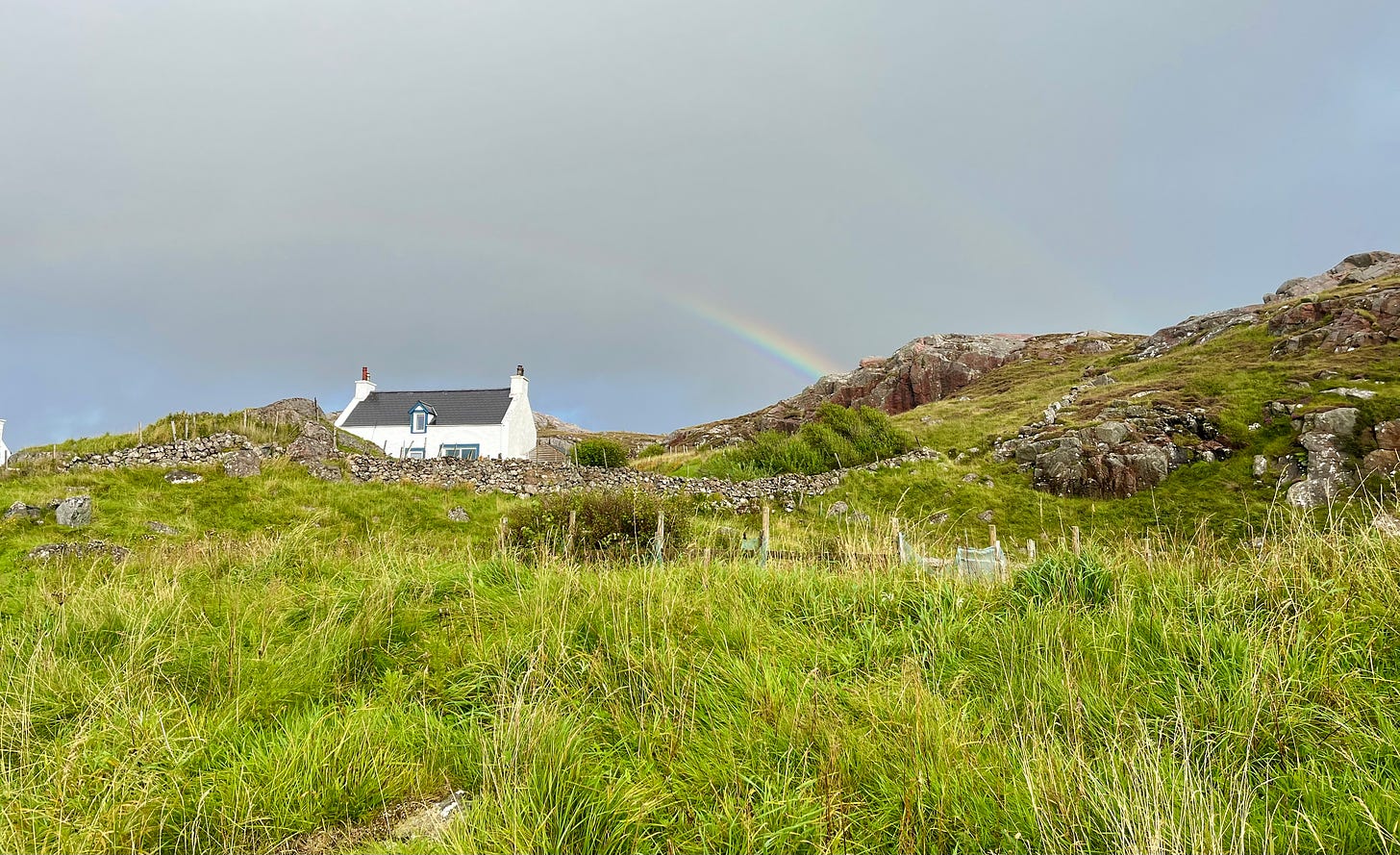 a white stone house on a rocky hilltop with course green pasture below and a dark gray sky cut by a rainbow above