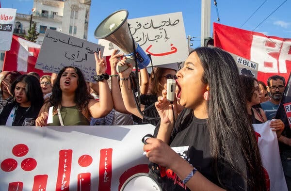 A woman shouts into a loudspeaker while demonstrators hold placards and carry a banner.
