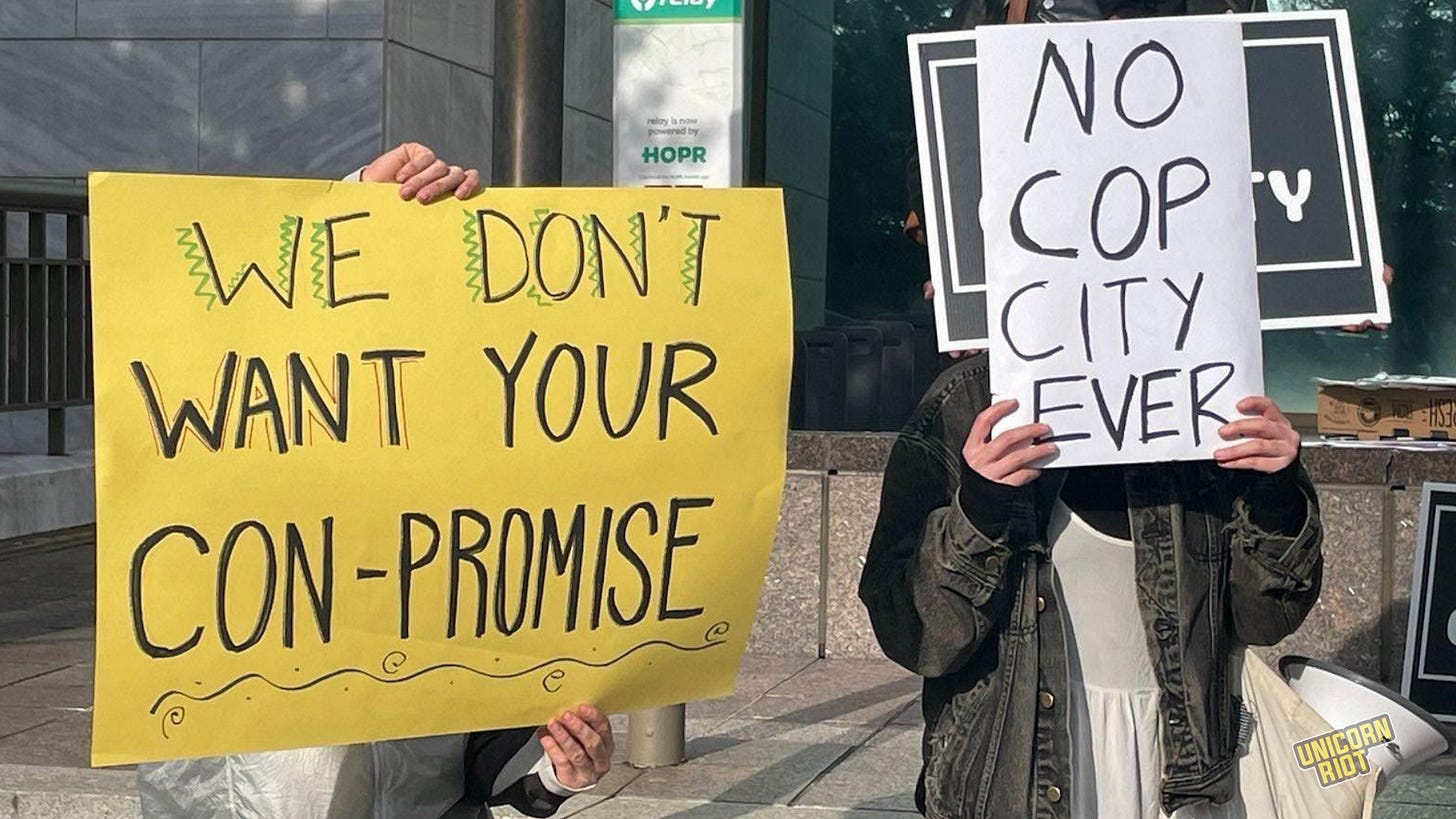 Two protesters hold signs covering their faces. One sign says "We don't want you con-promise" and the other says "no cop city ever."