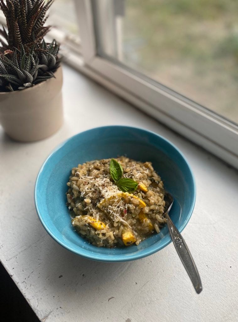 A blue bowl of pesto risotto, with pieces of roasted yellow zucchini throughout and grated parmesan on top. Two small basil leaves garnish the centre, and a fork sticks out at the front of the bowl. The bowl is set on a windowsill with a spiky plant in the background.