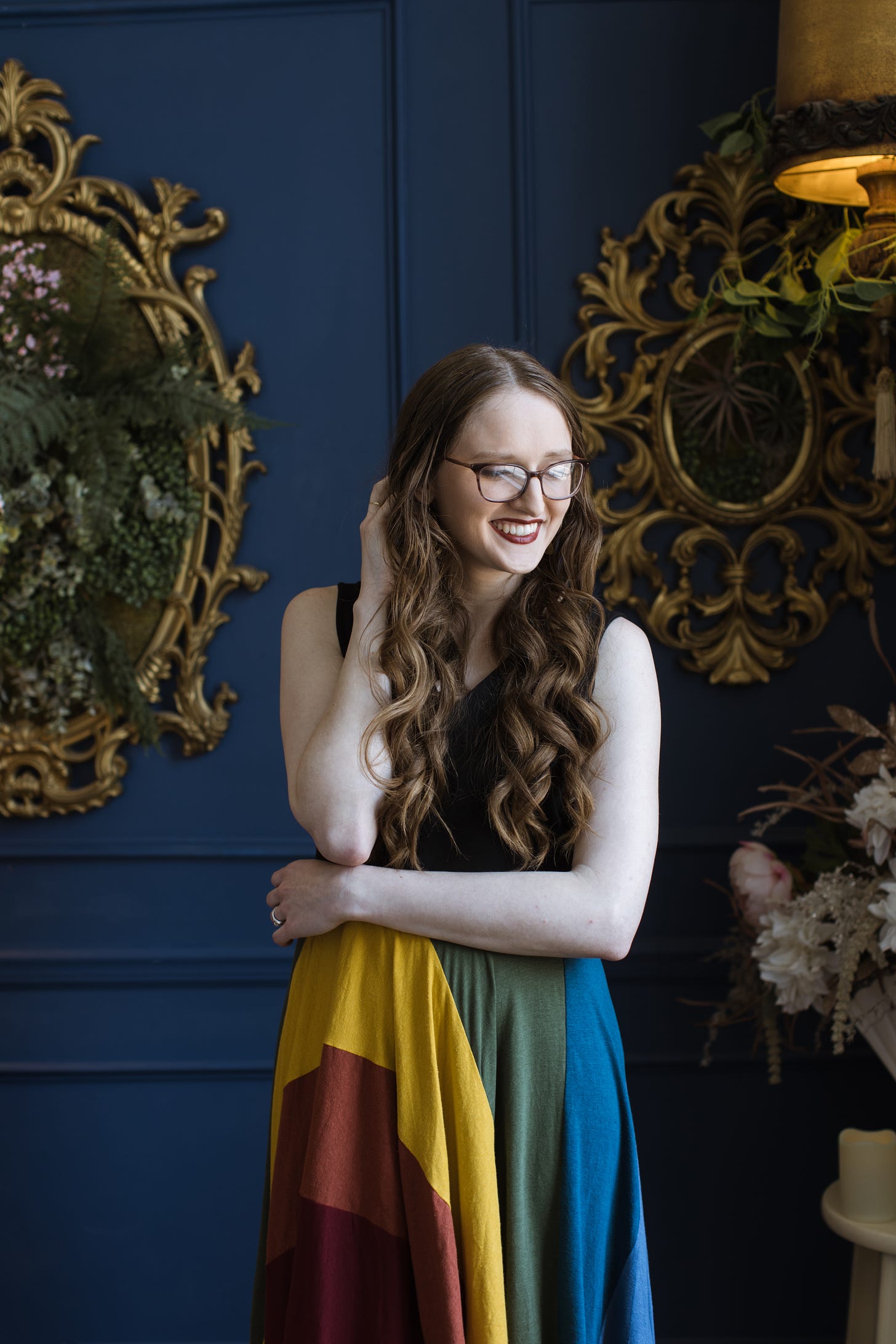 Kandi Zeller (she/her), a white woman with long, reddish brown hair and glasses, smiles and looks down. She is wearing a rainbow dress and is in front of blue backdrop with lots of green foliage.