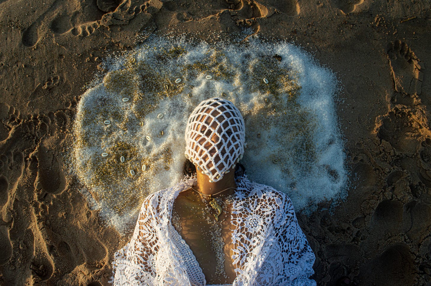 person wearing a white netted mask laced with cowry shells, the person is laying on the sand at a beach. Their head is surrounded by salt
