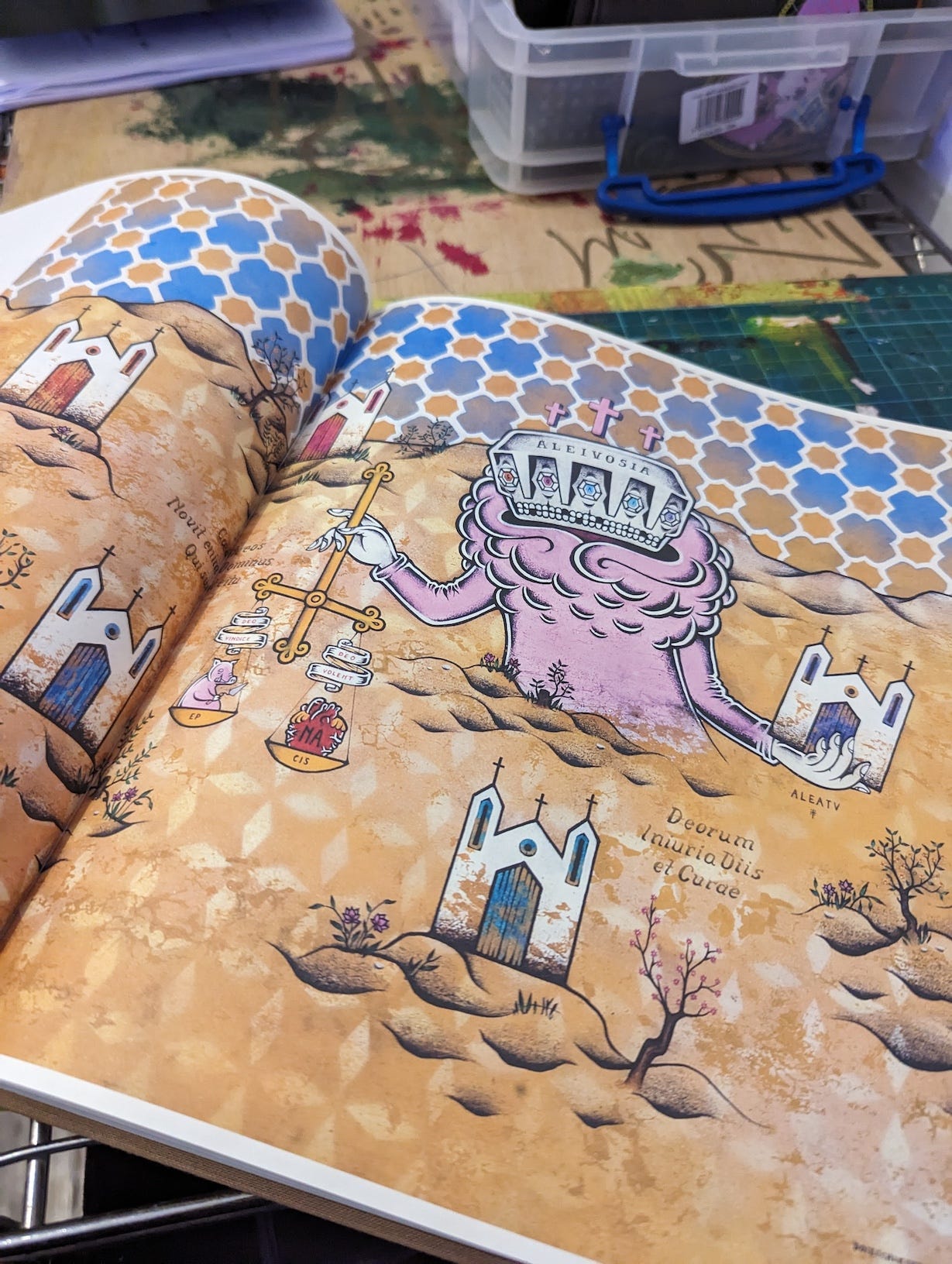 An interior page of Calma: The Art of Stephan Doitschinoff shows a colorful, surreal illustration. The scene includes a pink creature with a crown labeled "ALEIVOSIA" holding a golden scale. The background features a tiled pattern in shades of blue and orange, with small churches and barren landscapes scattered throughout. The artwork has a distinct, whimsical style, blending religious and mystical symbolism. The book is open on a workbench, showing the vivid, detailed illustrations.