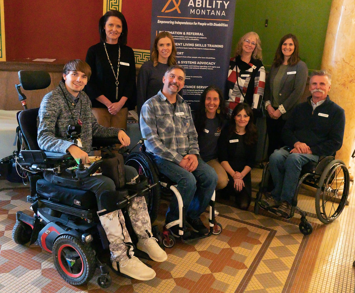 A group of smiling people pose in front of an Ability Montana banner -- three of them are in wheelchairs.