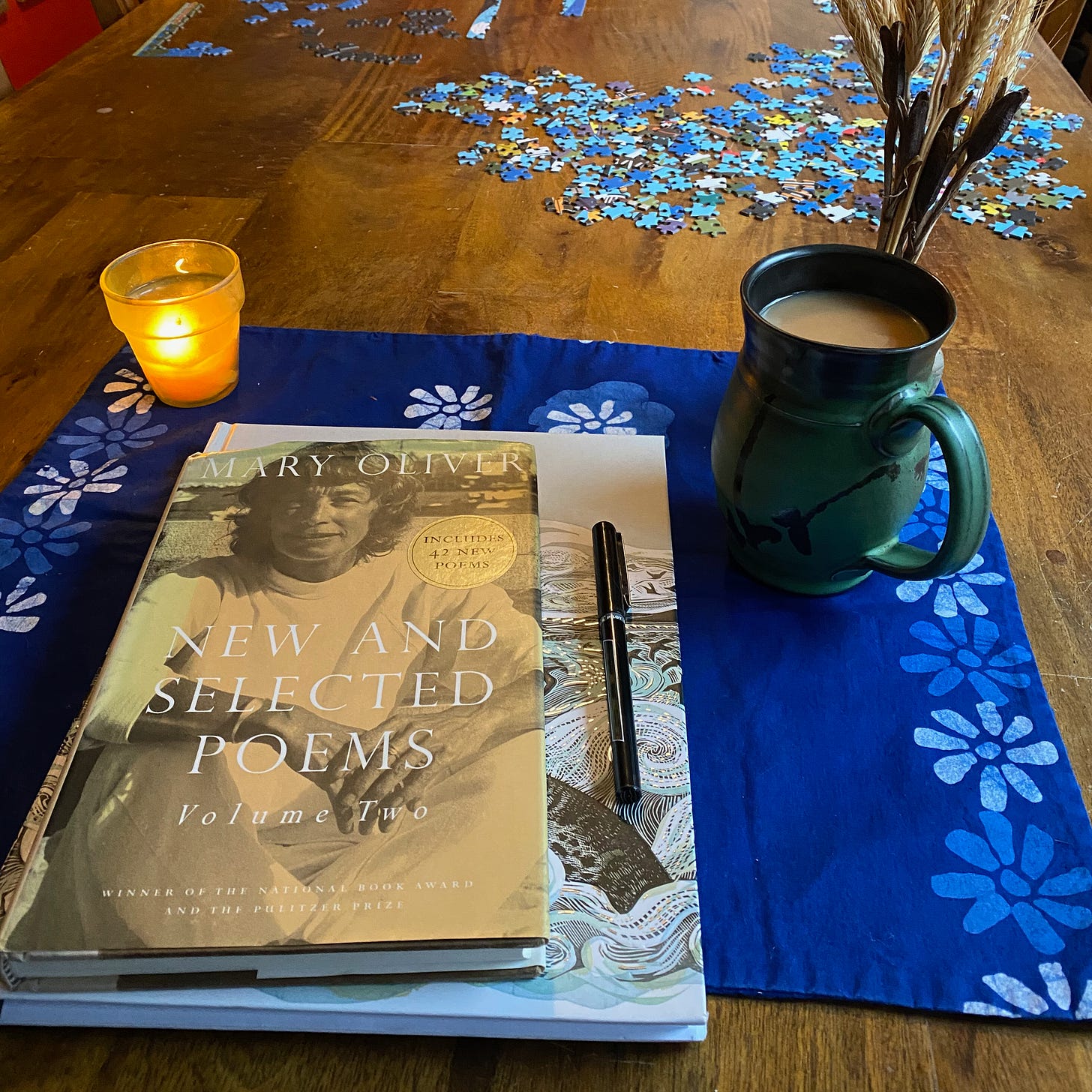 A notebook, pen, and book of Mary Oliver poems on a blue placemat next to a candle, mug of tea, and jar of dried grasses.