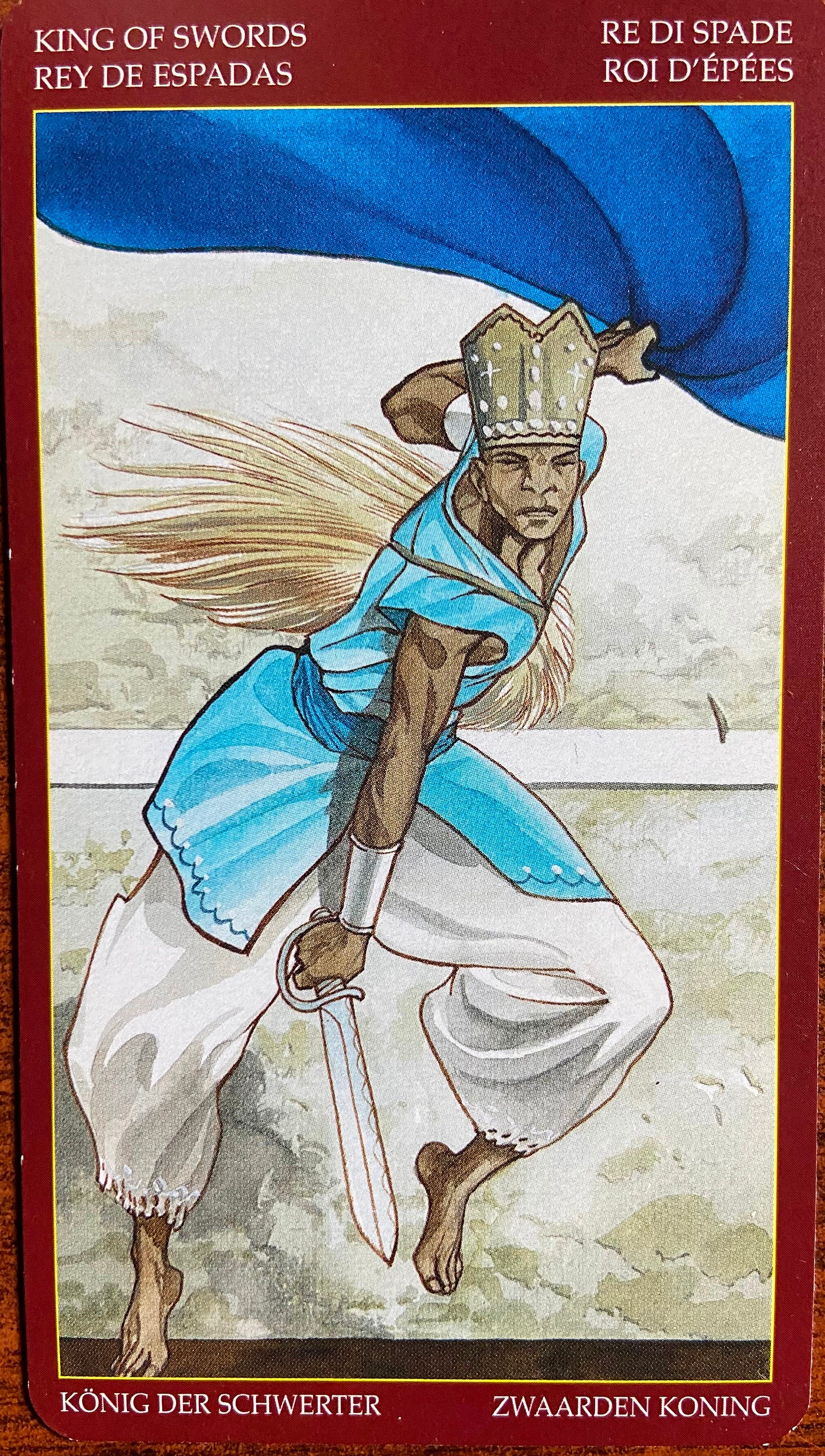 King of Swords: The barefooted, muscular figure is a Black man who spans most of the card’s space in a dynamic pose. Gazing inwards, face ahead, he raises a billowing blue fabric with his left hand while his right hand points a sword towards the ground. He wears an aqua-colored, sleeveless tunic over wide white pants and a mitre headdress which is a symbol of a bishop’s authority.