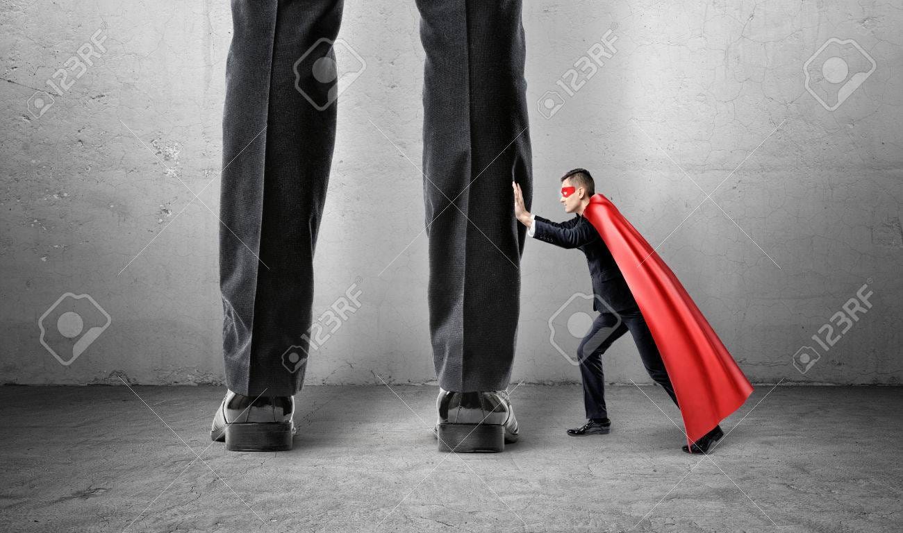 A small businessman in a  cape trying to push away giant man legs. - 77832325