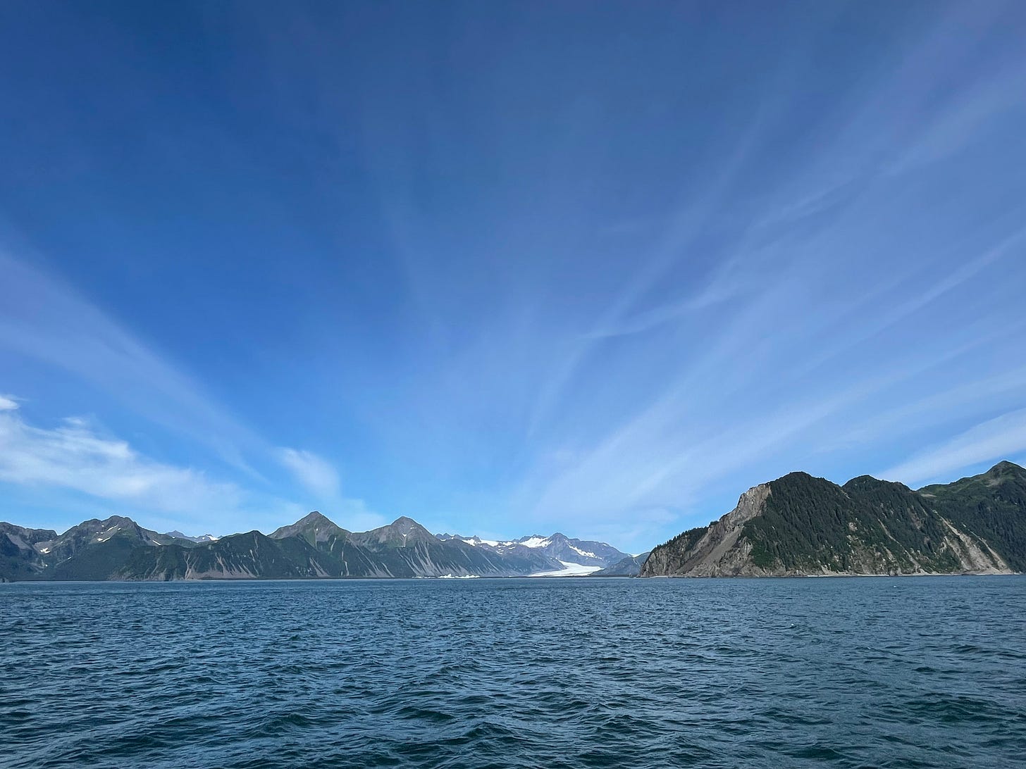 An Alaskan ford from a boat, snow-peaked mountains in the background and a blue sky with whispy streaks of cloud