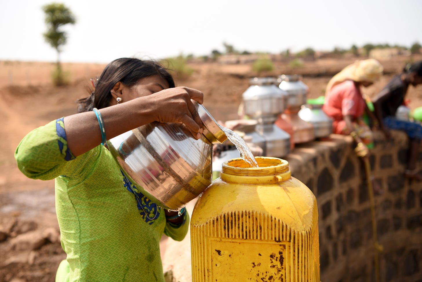 A woman in a green dress pours water from a silver pot into a yellow plastic container. Her arm and the pot obscure her face.