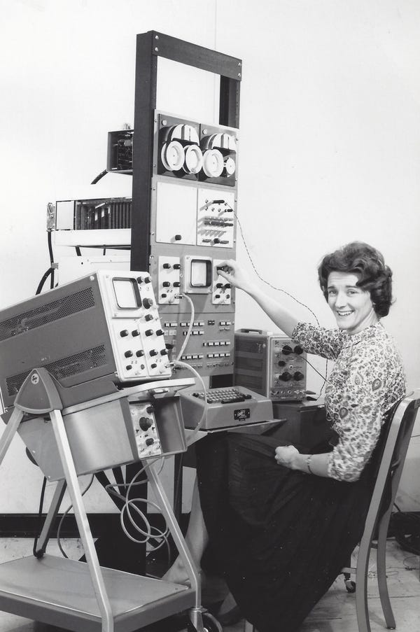 A historic photo of a woman happily working at a huge stack of old computer equipment.