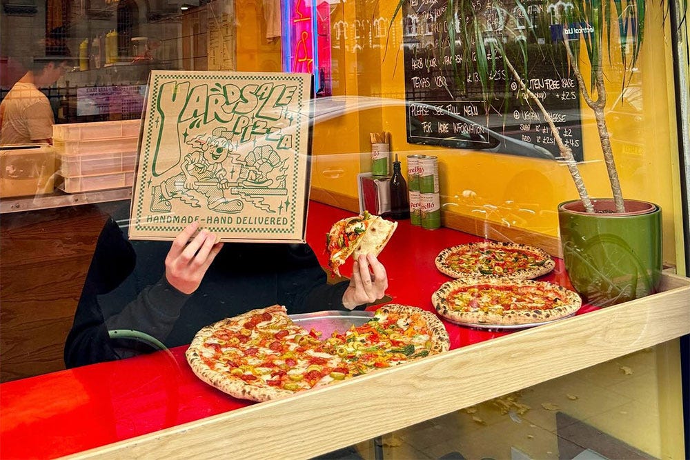 Person sitting in pizzeria window eating slice of pizza, holding a pizza box in front of their face as a disguise.