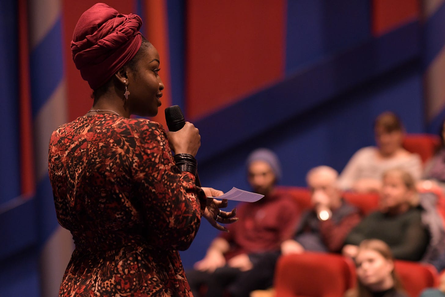 Lovette Jallow presenting in a red and blue auditorium to a seated audience, discussing autism, communication differences, and intersectional inclusion in educational and public settings. Lovette Jallow presenting in a red and blue auditorium to a seated audience, discussing autism, communication differences, and intersectional inclusion in educational and public settings.