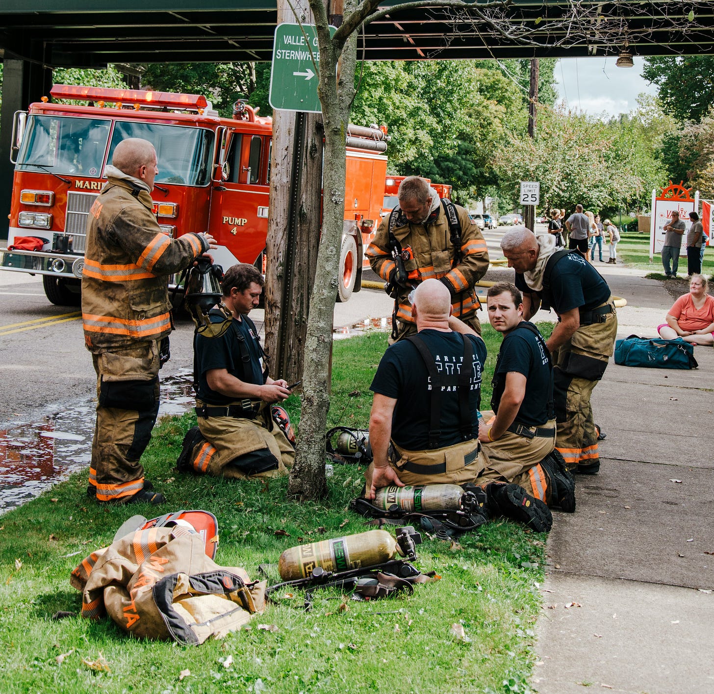 a group of firefighters in gear stand and kneel on a patch of grass next to a tree. a firetruck  is parked on the street next to them