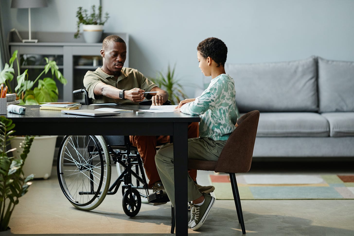 A black man in a manual wheelchair is sitting at a table with a younger boy. It could be a school or an office .