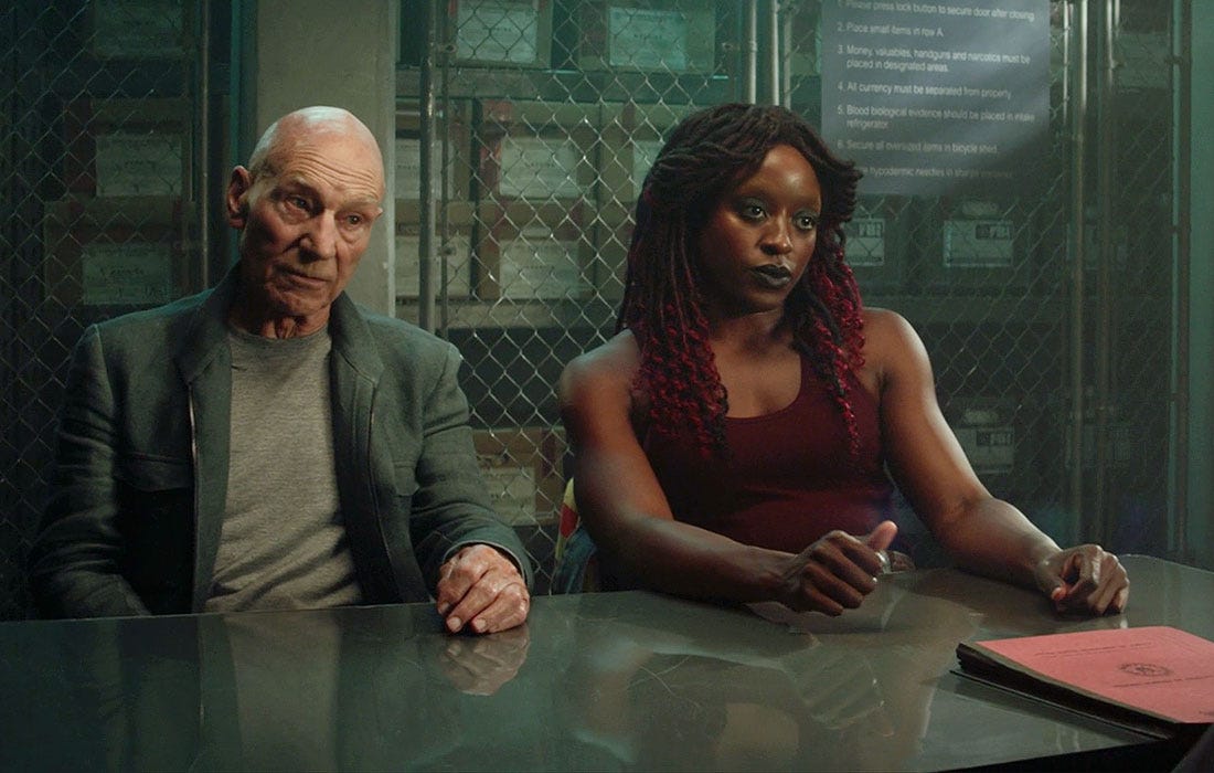 This image from "Mercy" (Star Trek: Picard Season 2's eighth episode) shows Jean-Luc Picard (played by Patrick Stewart, left) and Young Guinan (played by Ito Aghayere, right) seated at a table inside a basement FBI interrogation room. Both characters gaze dubiously at Jay Karnes's offscreen Agent Martin Wells. An FBI dossier lies on the table in front of Wells. This image from "Mercy" (Star Trek: Picard Season 2's eighth episode) shows Jean-Luc Picard (played by Patrick Stewart, left) and Young Guinan (played by Ito Aghayere, right) seated at a table inside a basement FBI interrogation room. Both characters gaze dubiously at Jay Karnes's offscreen Agent Martin Wells. An FBI dossier lies on the table in front of Wells.