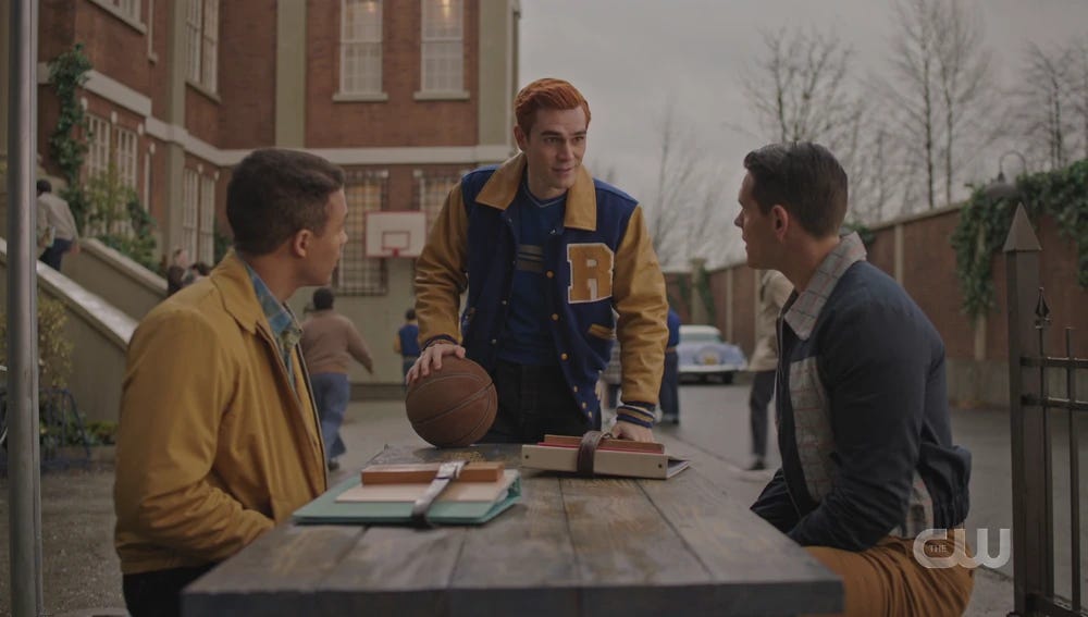 Archie standing at the head of a table talking to Kevin and Clay sitting on either side of the table.