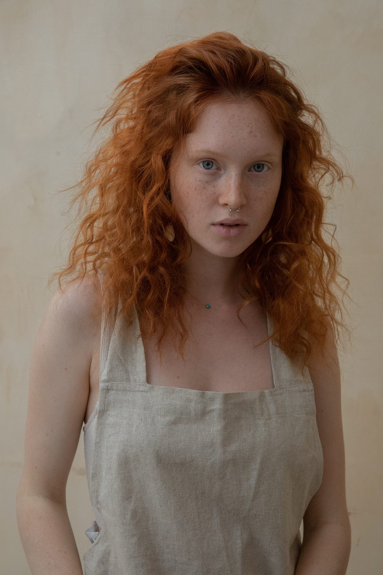 A young woman with curly, red hair, fair skin, and freckles wears an apron. She looks at the camera with a neutral expression.