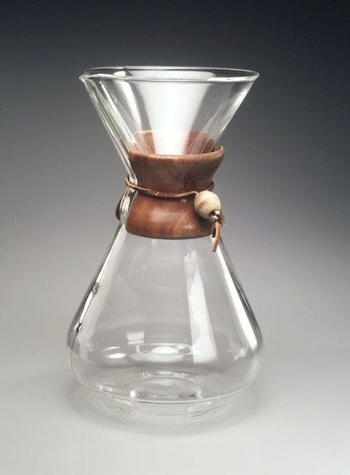 A close-up of a Chemex coffee brewer, a glass bulb with a flat bottom that pinches in the middle with a glass cone and spout at the top. A wooden collar wraps around the pinched middle portion to protect the hand from heat and provide grip for pouring.