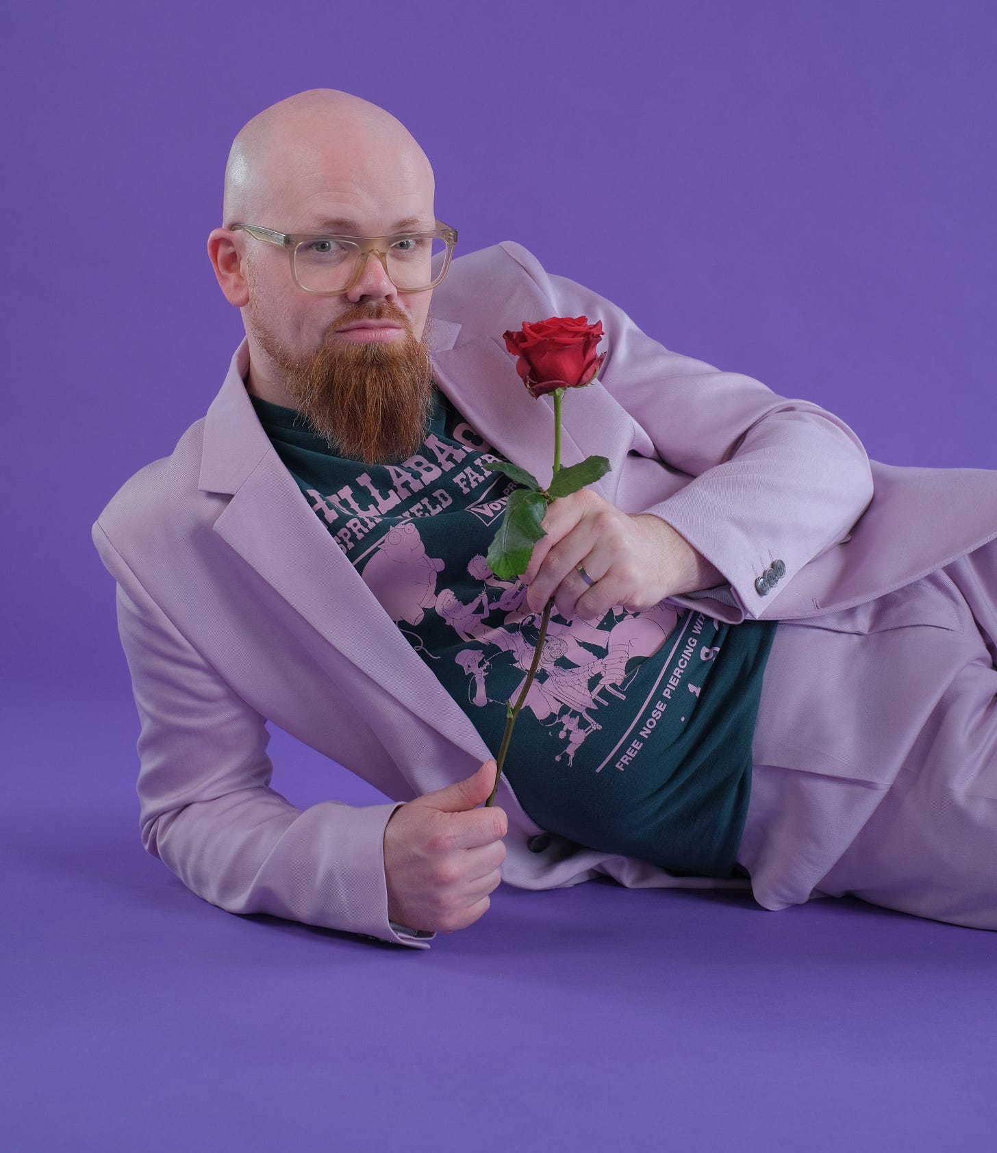 A man wearing a purple suit lays on the floor, holding a rose out to the viewer. He has a shaved head, wears glasses, and sports a brown beard.