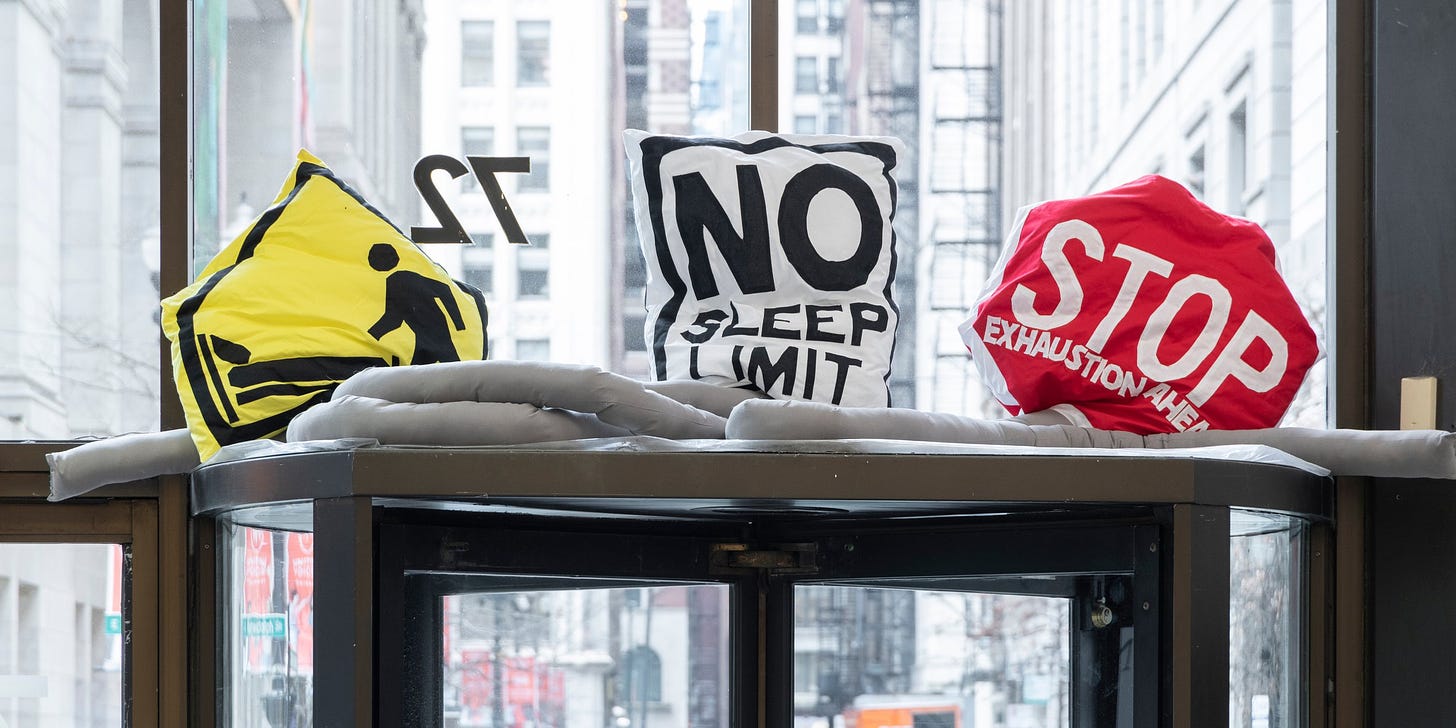 A photograph of pillows that look like road signs with modified text: STOP EXHAUSTION AHEAD. NO SLEEP LIMIT. They are positioned on top of a revolving door.