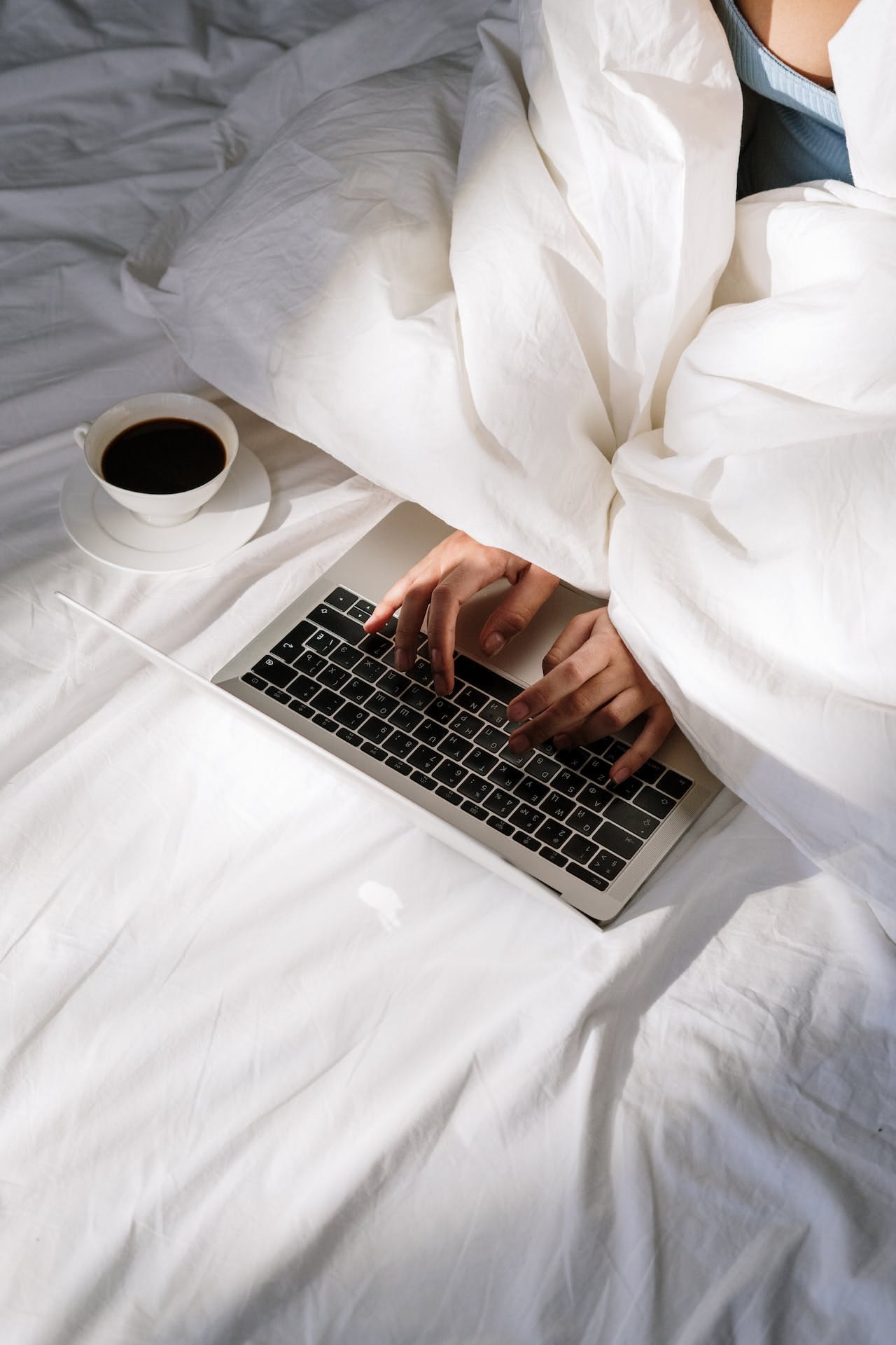 person typing on computer while under sheets of bed with coffee