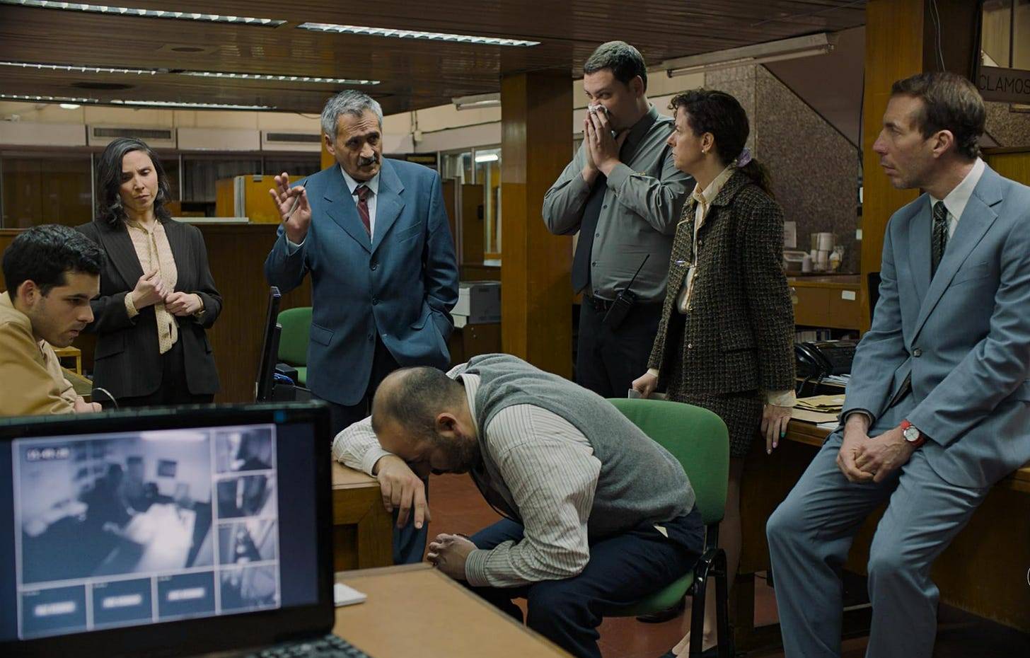 Several men and women dressed in business attire listen as an older man, who is making a gesture with his thumb and forefinger, speaks to them in a woodpanelled office setting.