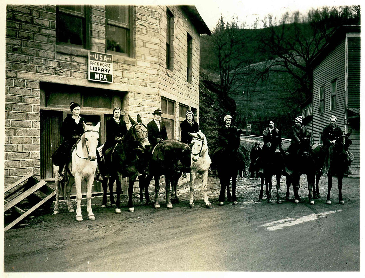 Eight women in warm clothing sit astride horses on a street. A sign behind them says USA Pack Horse Library WPA. The image is black and white, and is from 1938