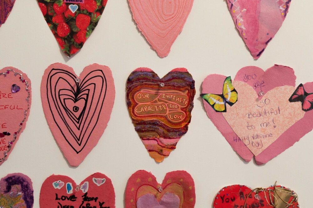 A closeup of a white wall covered in handmade pink valentine cards in the shape of hears. One of them says, out infinite capacity for love.