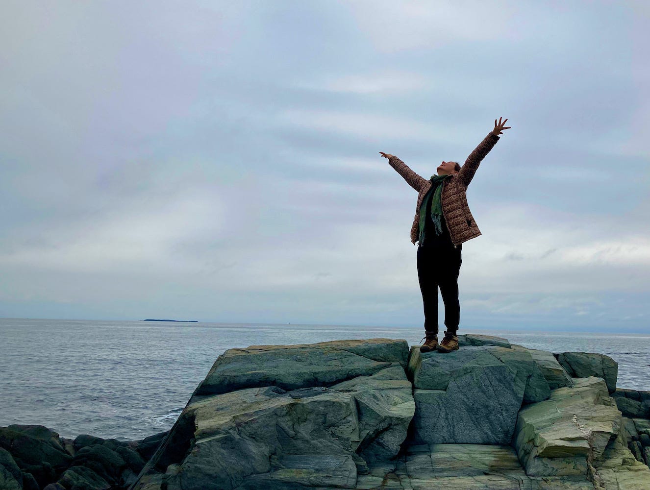  Tamiko Beyer stands on rocks in front of a calm oceanscape, arms spread wide, face lifted to the soft gray sky