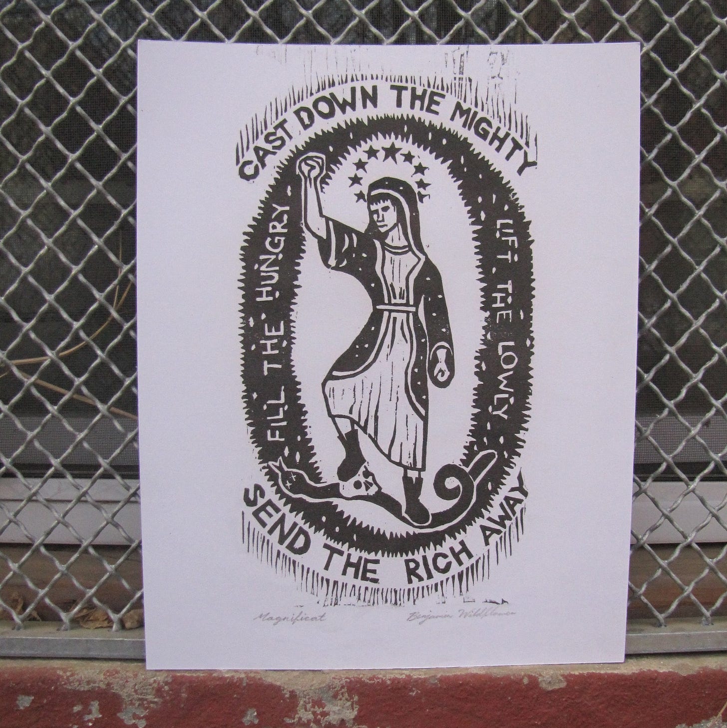 A print hung against a chain link fence showing Mary trampling a snake with the text Cast Down the Mighty, Send the Rich Away. Fill the Hungry, Lift the Lowly.