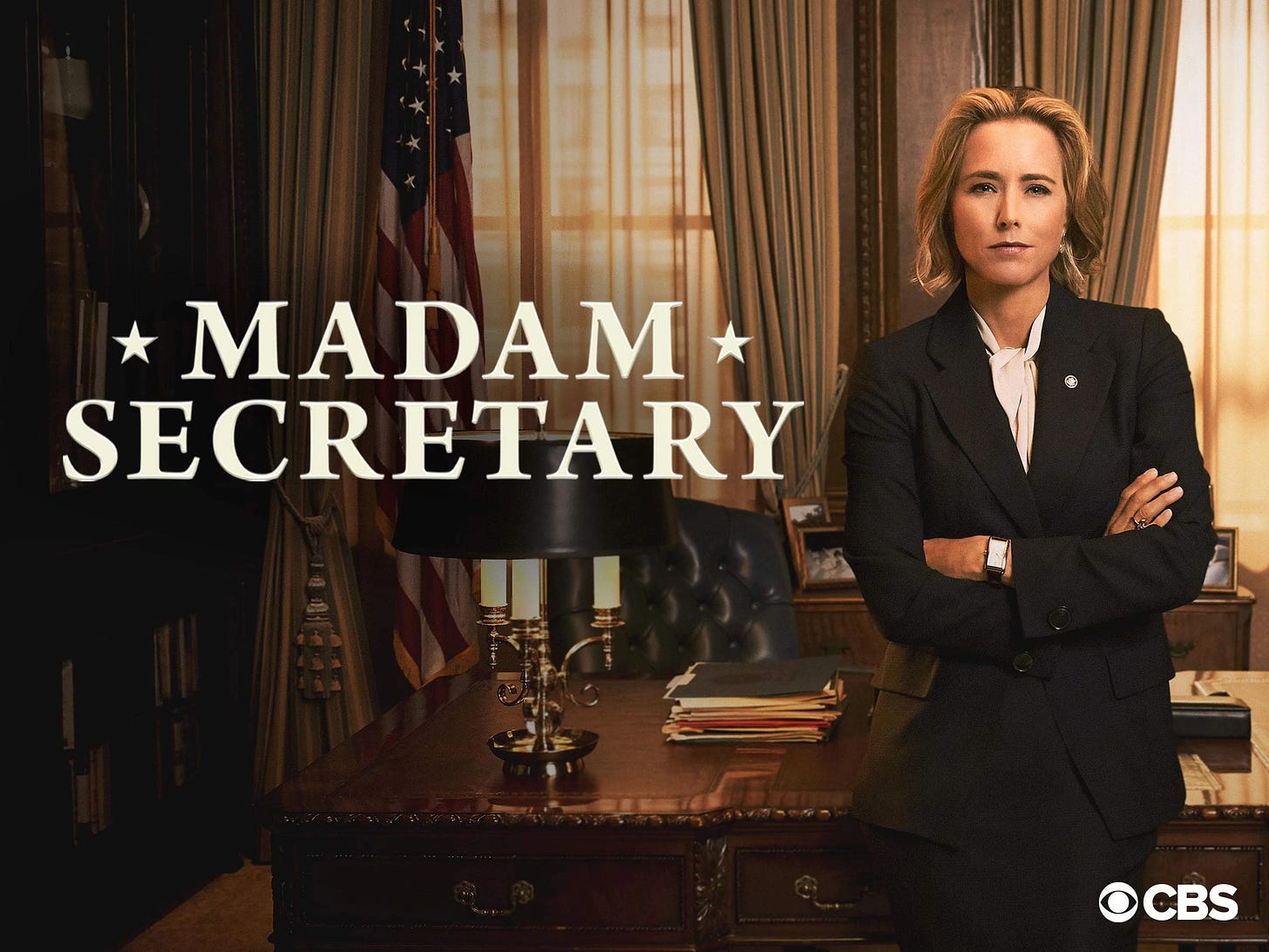 An image of Téa Leoni in her role as Madam Secretary. Arms folded, in a suit, leaning on an official looking desk. 