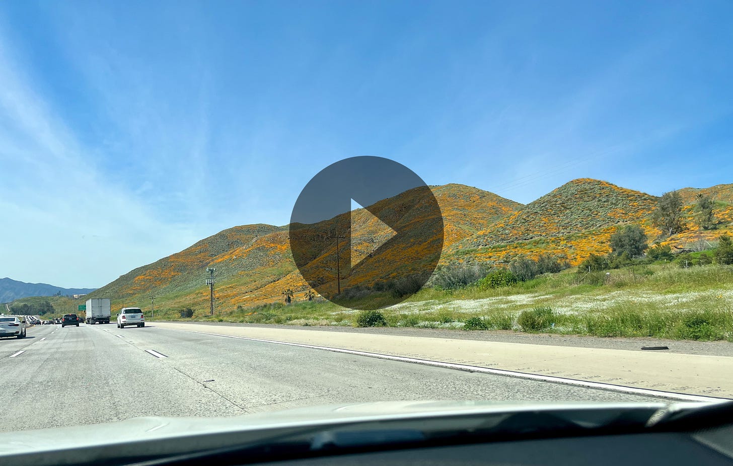 A photo with a play button on top of it. The play button is taken from the passenger seat of a car driving on the freeway. There's a road ahead and, to the side, hills covered in yellow poppy flowers. The blue sky is above.