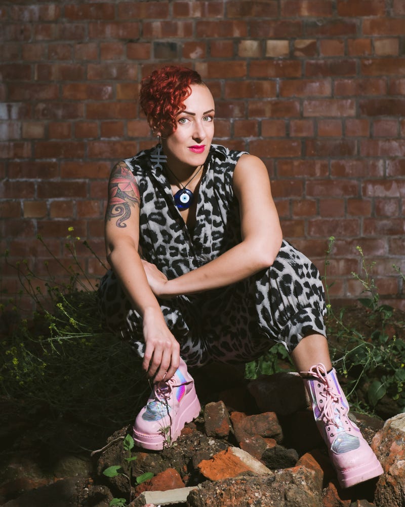 Woman with vivid red curly hair, tattoos and pink plaforms crouched and looking dead cool on some bricks. Jane Claire Bradley is wearing signature leopard print!