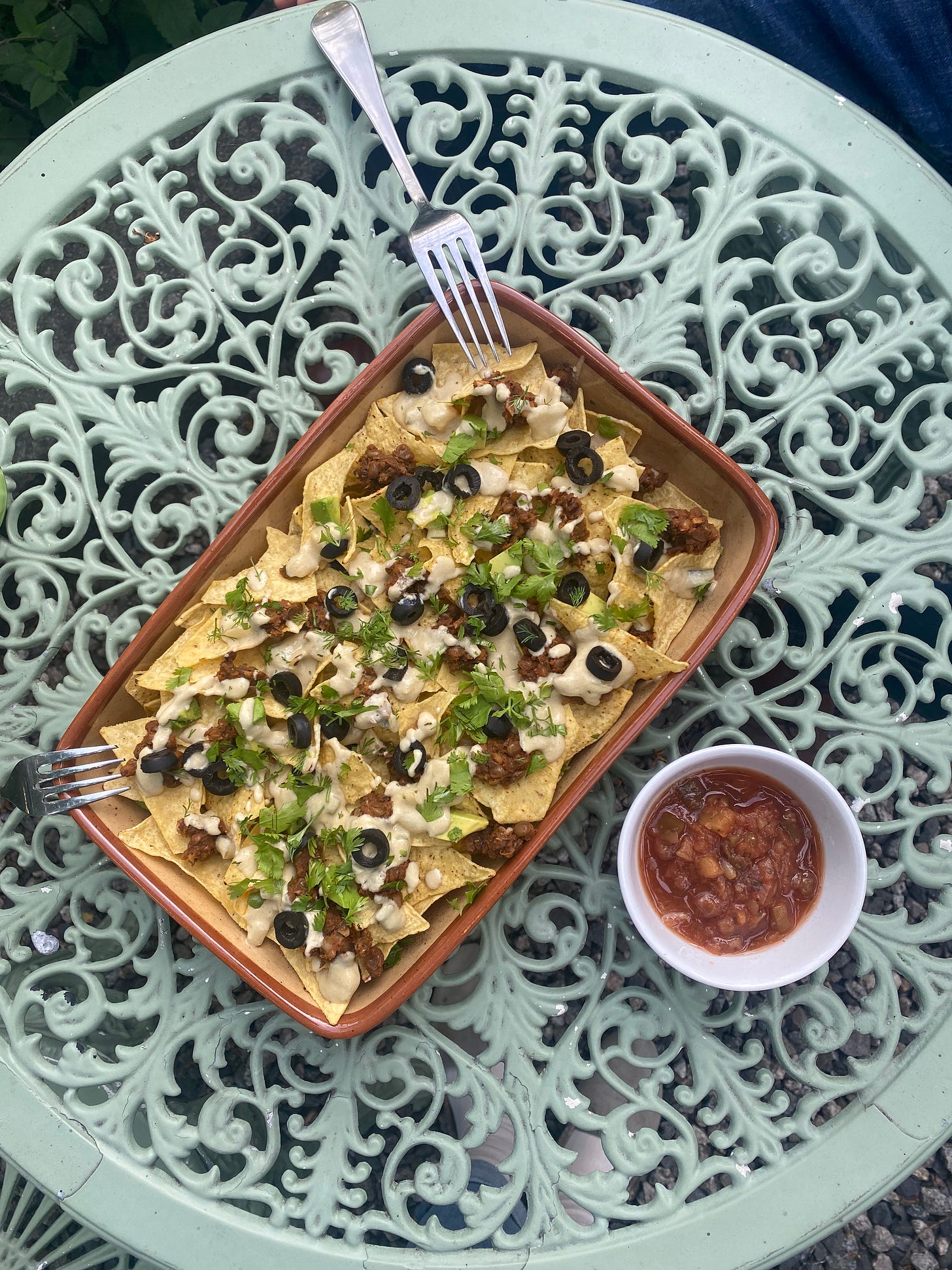 On a mint green outdoor café table, a rectangular stoneware dish of the nachos described above: queso, black olives, lentil meat, and cilantro spread over top. A small white dish of red salsa sits to the side.