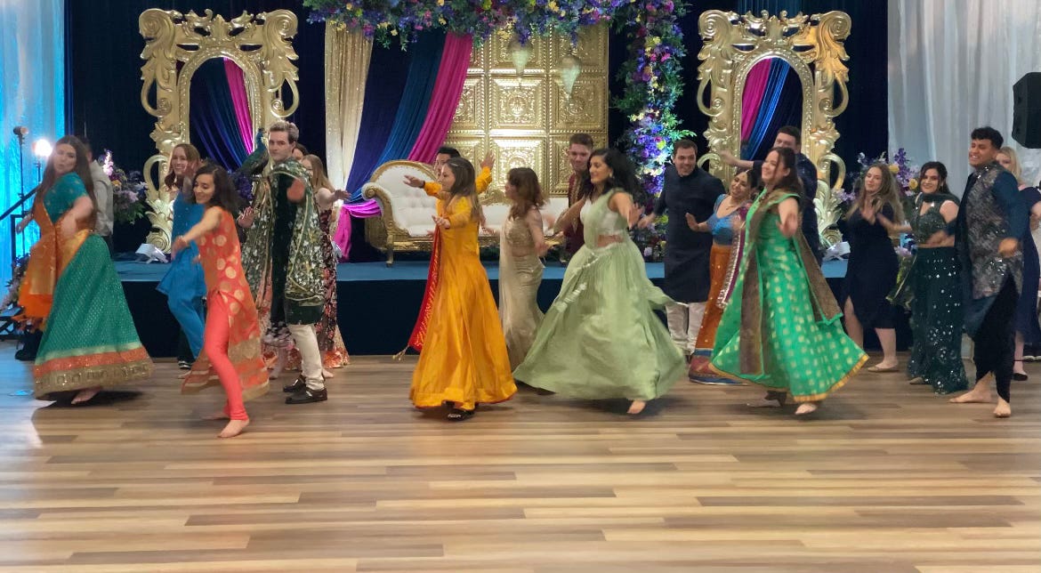 A group of men and women, in their 20s and 30s, dances wearing traditional Indian attire