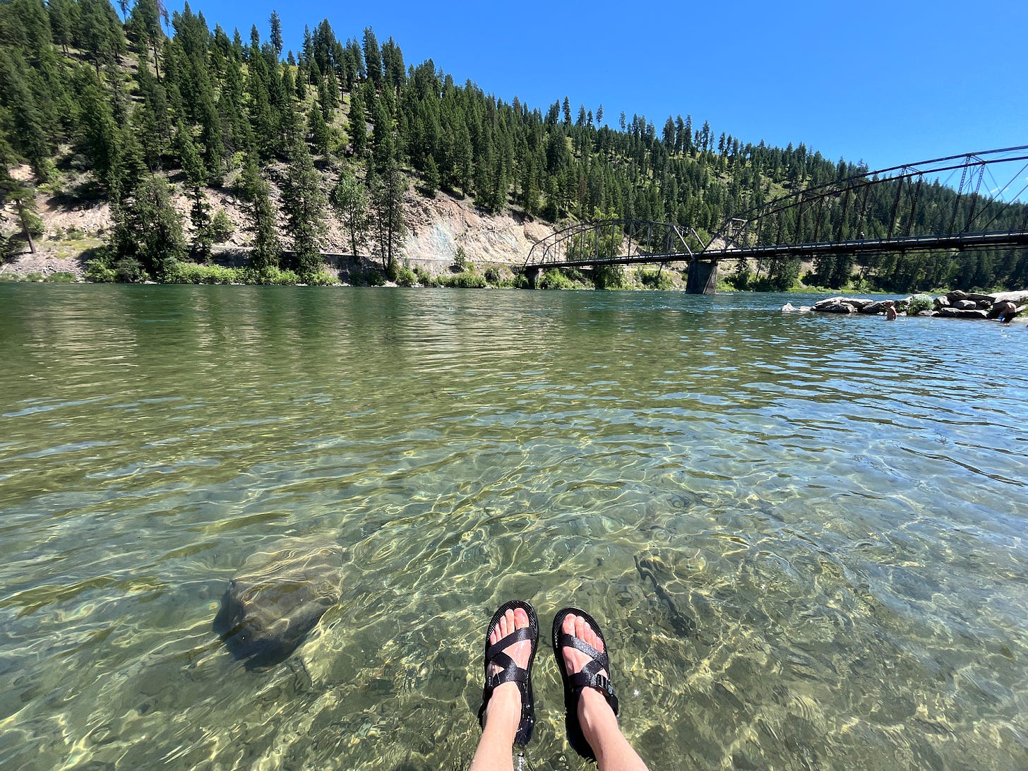 A mountain river with forest in the background two feet in sandals at the bottom of the frame.