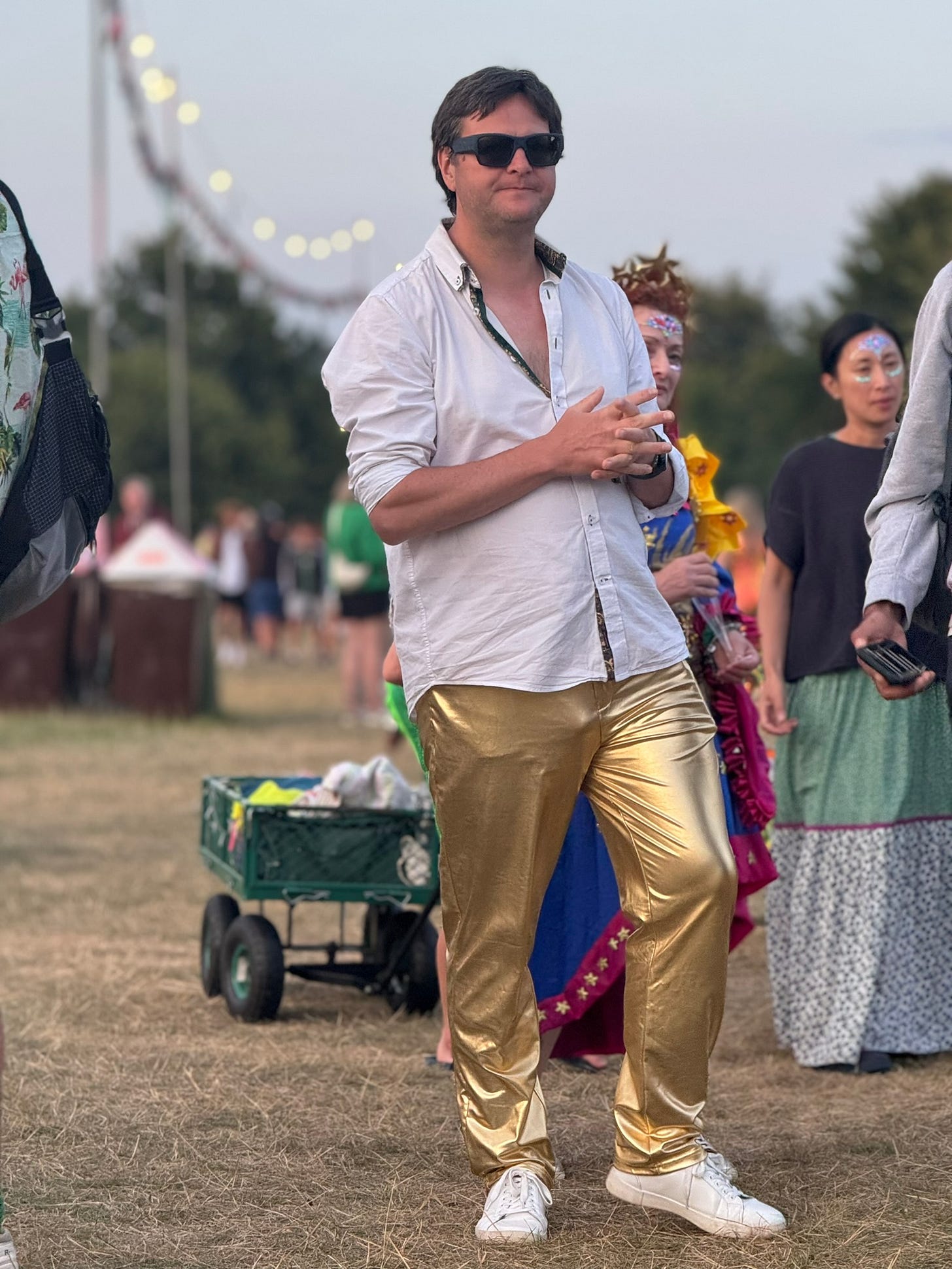 Man has brown hair, sunglasses, white shirt and gold shimmery trousers.  There are other people dressed up around him. 