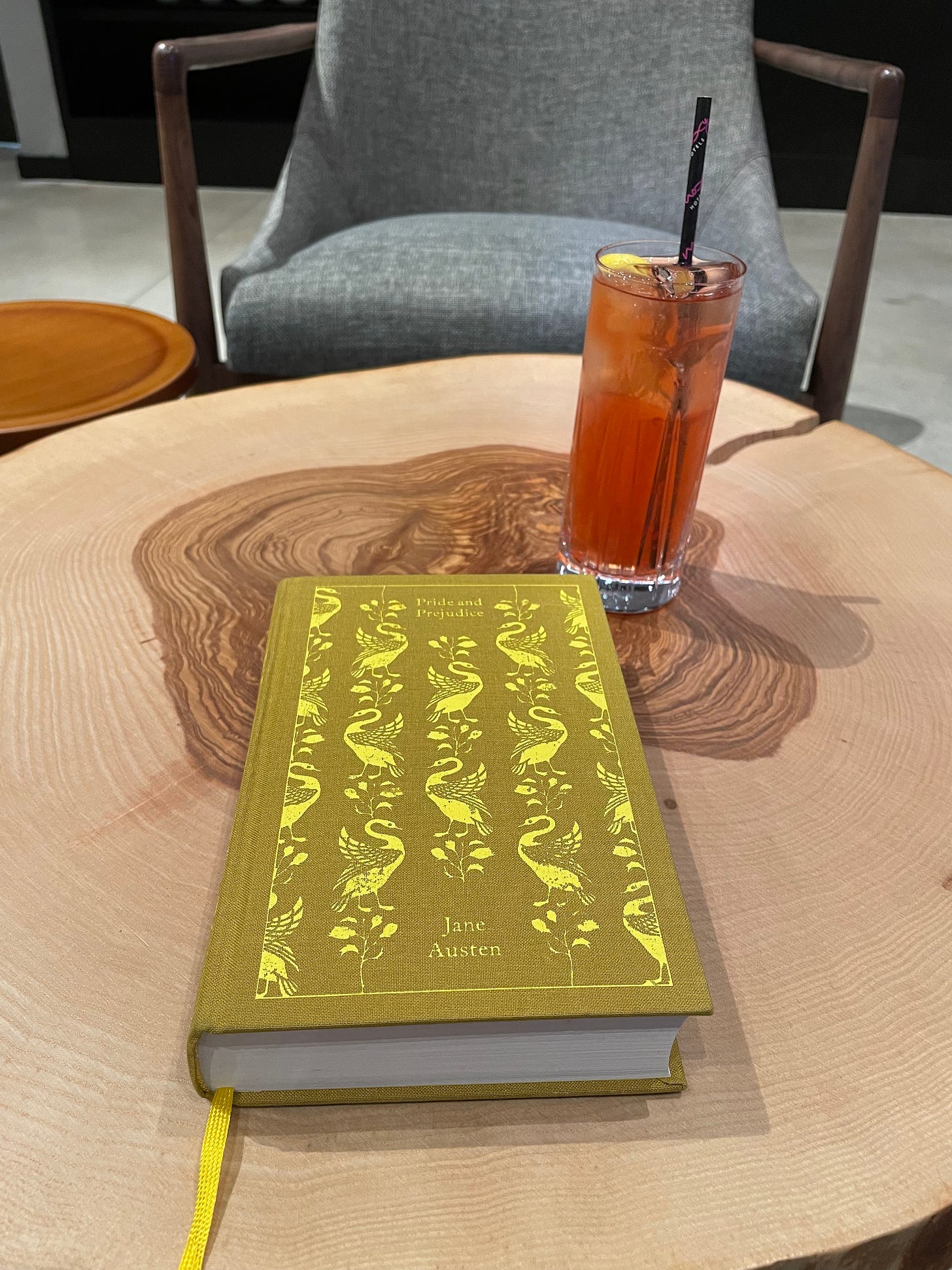 A clothbound version of Pride and Prejudice ona. table with an orange drink and chair in the background. 