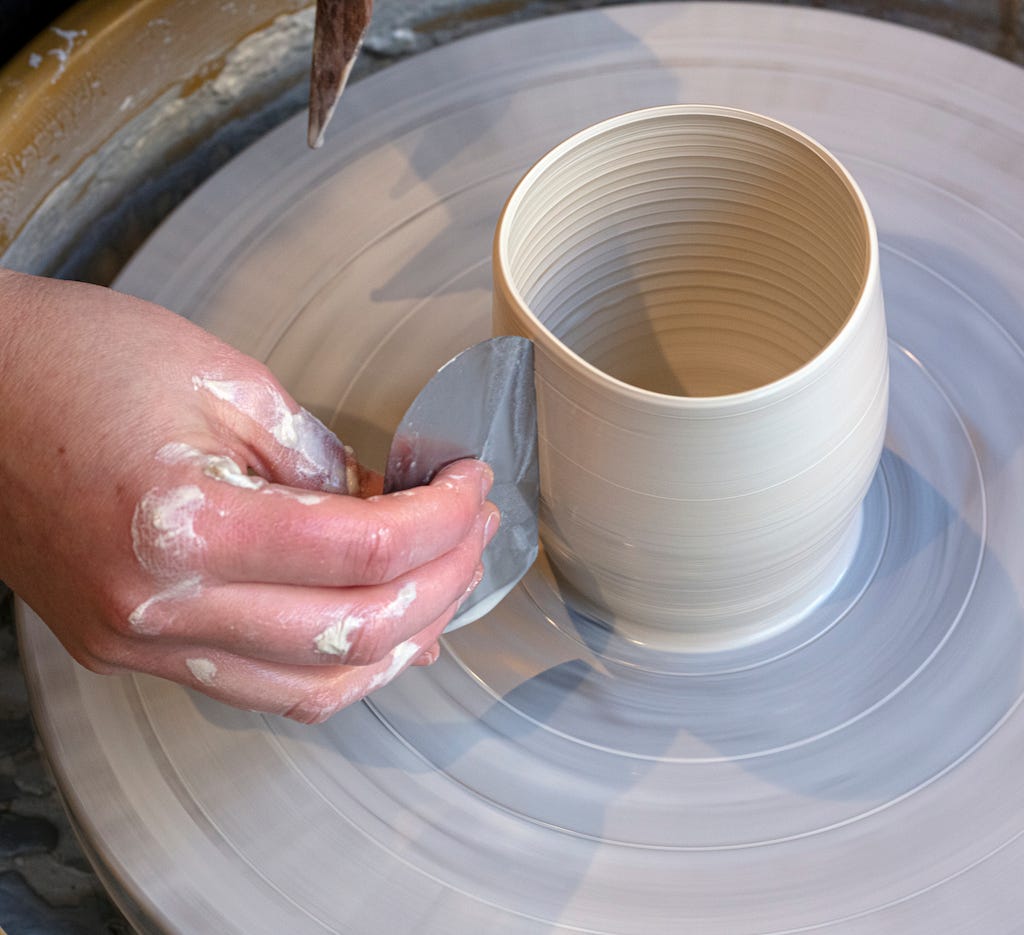 Right hand scraping side of porcelain cup on wheel with a metal rib. left hand, just out of sight, is holding a wooden throwing stick with a sharp point, to clean up the wheel head