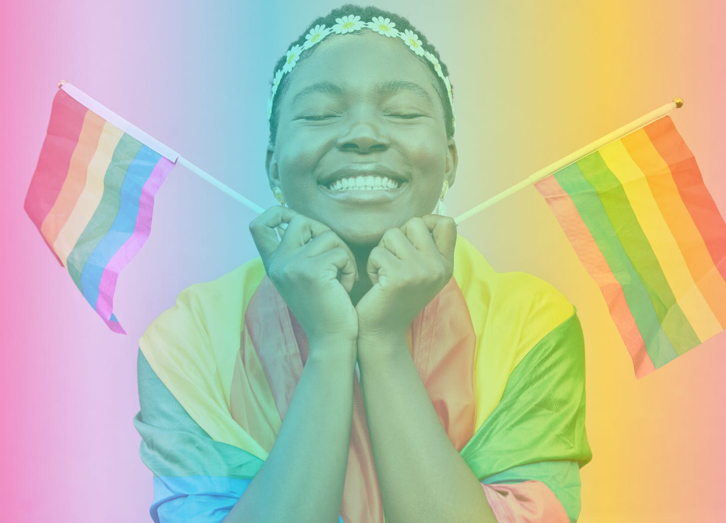 A rainbow-filtered image of a Black femme wrapped in a pride flag and holding two mini pride flags in their hands. They are smiling and have a headband on made of plastic daisies.